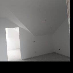 USŁUGI OGÓLNOBUDOWLANE - KAMIL KOBIELA - Wnętrze poddasza po malowaniu na biało, widoczne gniazdka elektryczne, surowy beton na podłodze i wystające kable. Otwór drzwiowy prowadzi do kolejnego pomieszczenia.