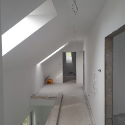 USŁUGI OGÓLNOBUDOWLANE - KAMIL KOBIELA - Poddasze po malowaniu na biało, widoczne okno dachowe, surowa ościeżnica i wystające przewody elektryczne. Na podłodze stoi wiadro farby.