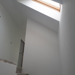 USŁUGI OGÓLNOBUDOWLANE - KAMIL KOBIELA - Wnętrze poddasza po malowaniu na biało, widoczne schody w trakcie wykończenia i okno dachowe z drewnianą ramą. Minimalistyczny design, surowe wykończenie.