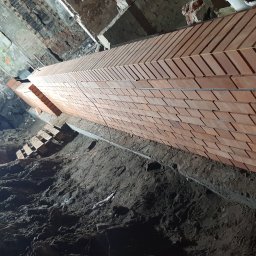 USŁUGI OGÓLNOBUDOWLANE - KAMIL KOBIELA - Wznoszenie muru z cegły klinkierowej w wykopie fundamentowym, widoczne zbrojenie i fragment rusztowania. Ujęcie z dołu, podkreślające pionowość konstrukcji.