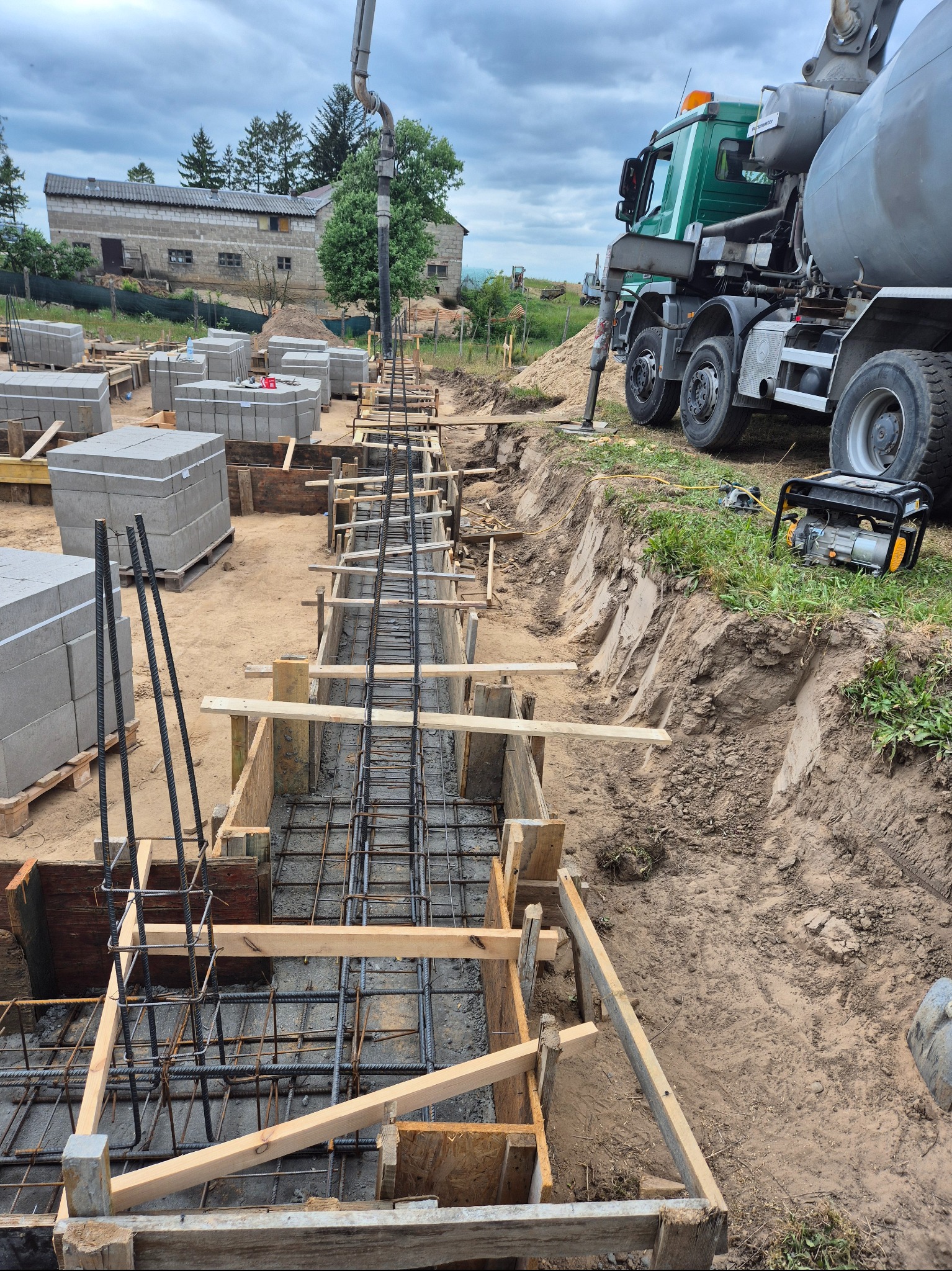 Zbrojenie fundamentów w drewnianym szalunku, widok z góry. Na drugim planie betonowóz i budynek gospodarczy. Etap budowy domu z użyciem betonu i stali.