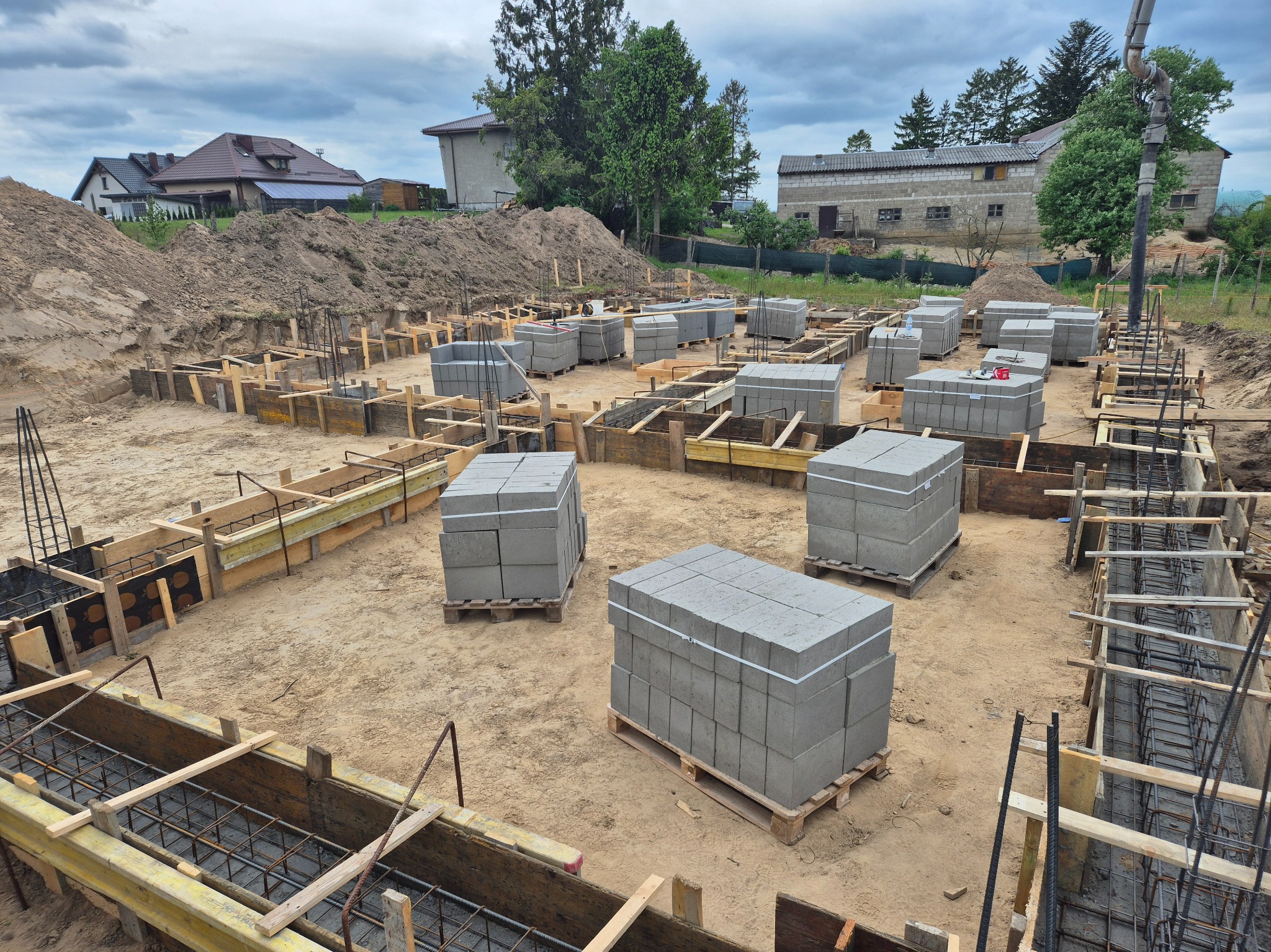 Fundamenty domu w trakcie budowy: szalunki zbrojeniowe, bloczki betonowe na paletach, ziemia. W tle budynki mieszkalne i drzewa pod pochmurnym niebem.