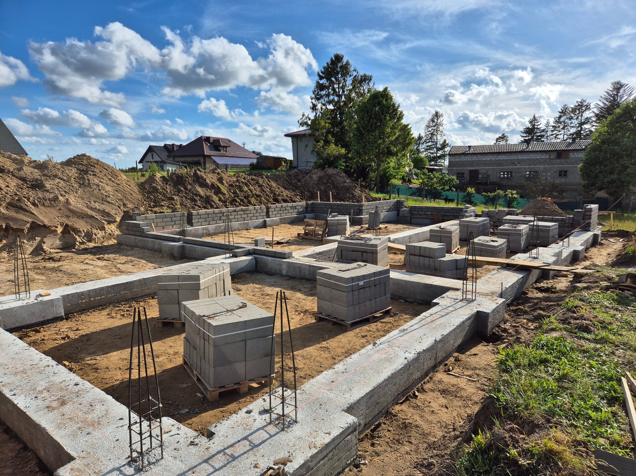 Fundamenty domu w trakcie budowy, zbrojenie wystaje, bloczki betonowe na paletach, w tle domy i drzewa pod błękitnym niebem z chmurami.