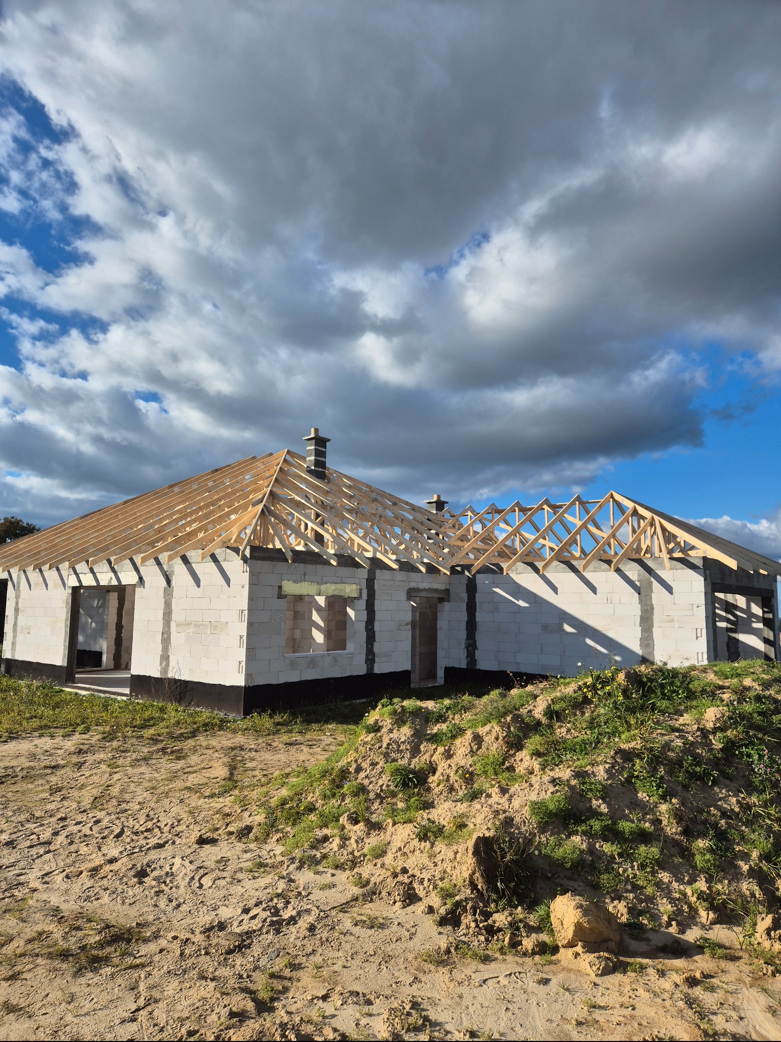 Surowy dom z białych bloczków z drewnianą konstrukcją dachu w trakcie budowy, na tle pochmurnego nieba. Widoczny fragment terenu z piaskiem i roślinnością.