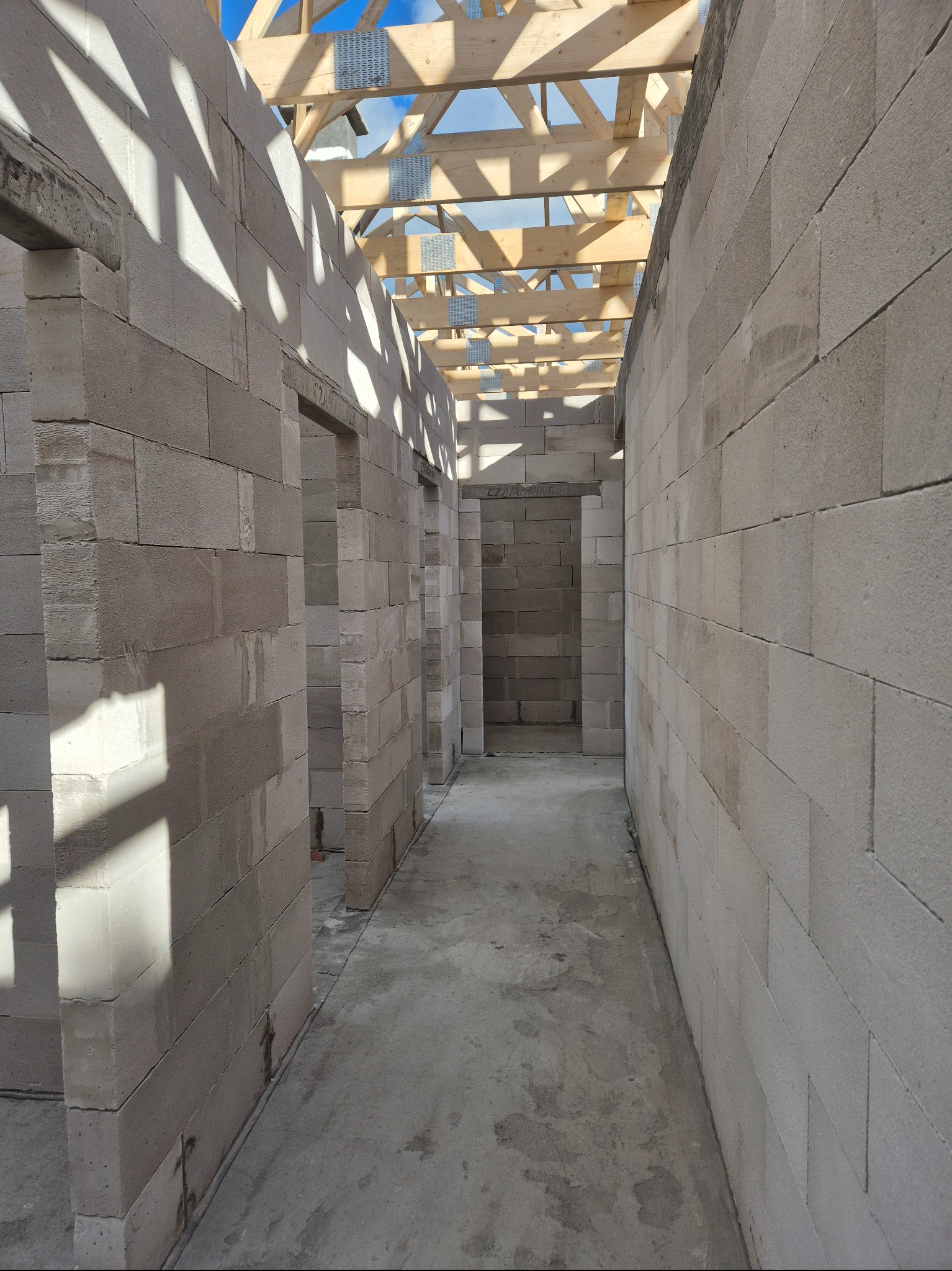 Surowy stan wnętrza domu z bloczków, widoczne ściany działowe i drewniana konstrukcja dachu z metalowymi łącznikami, betonowa podłoga, wpadające światło słoneczne.