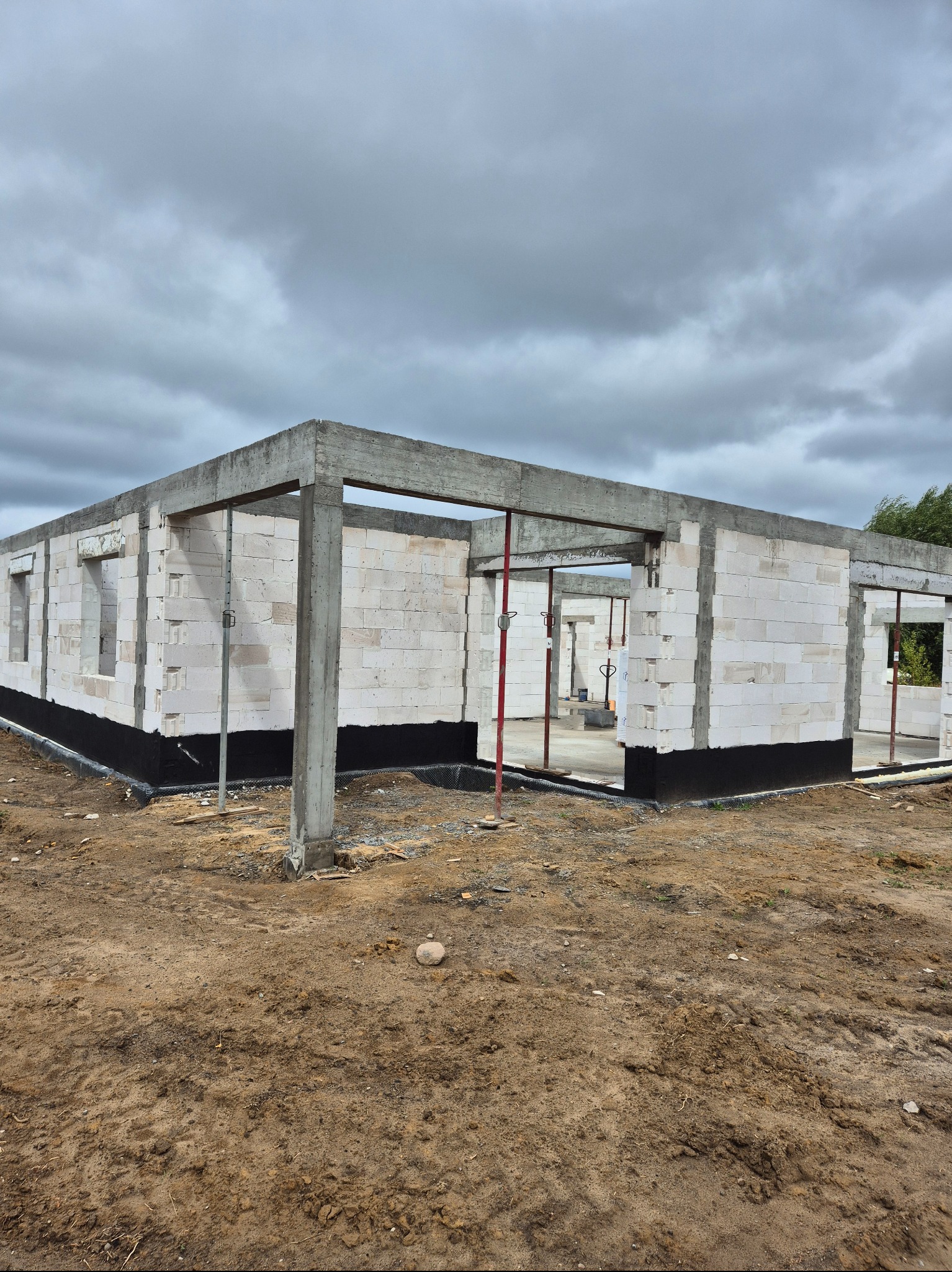 Surowy stan domu z betonu komórkowego w trakcie budowy, widoczne podpory i izolacja fundamentów, pochmurne niebo w tle. Ujęcie z poziomu gruntu.