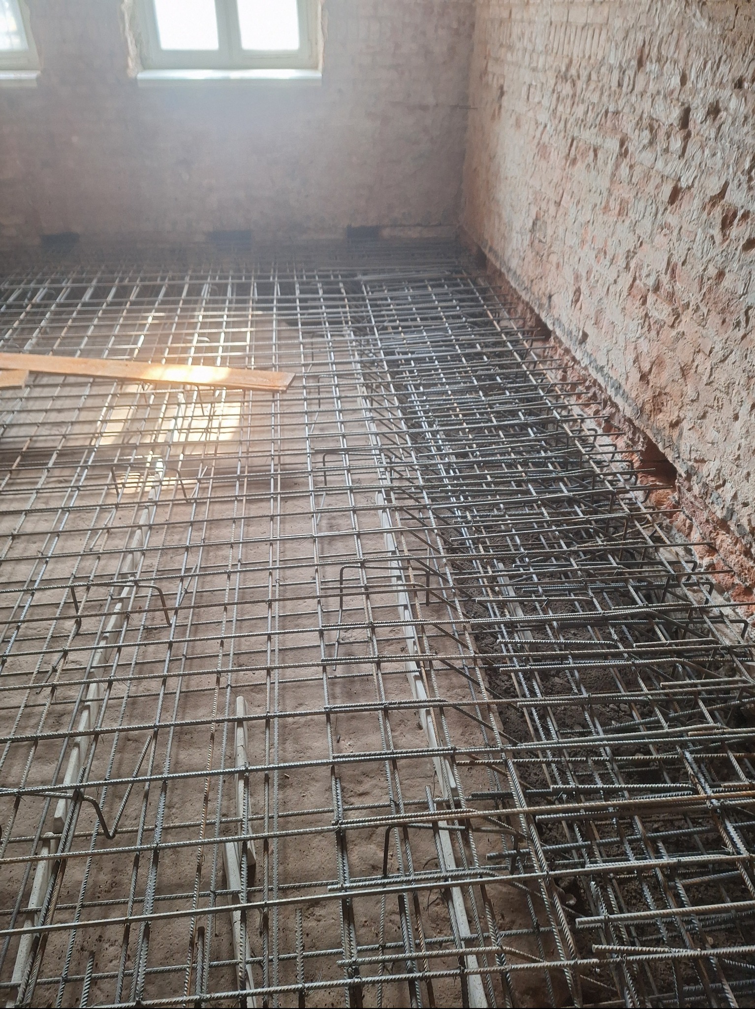 Zbrojenie pod wylewkę na tle ceglanej ściany i okna. Widoczne metalowe pręty tworzące siatkę zbrojeniową, przygotowane do zalania betonem. Surowy stan wnętrza.