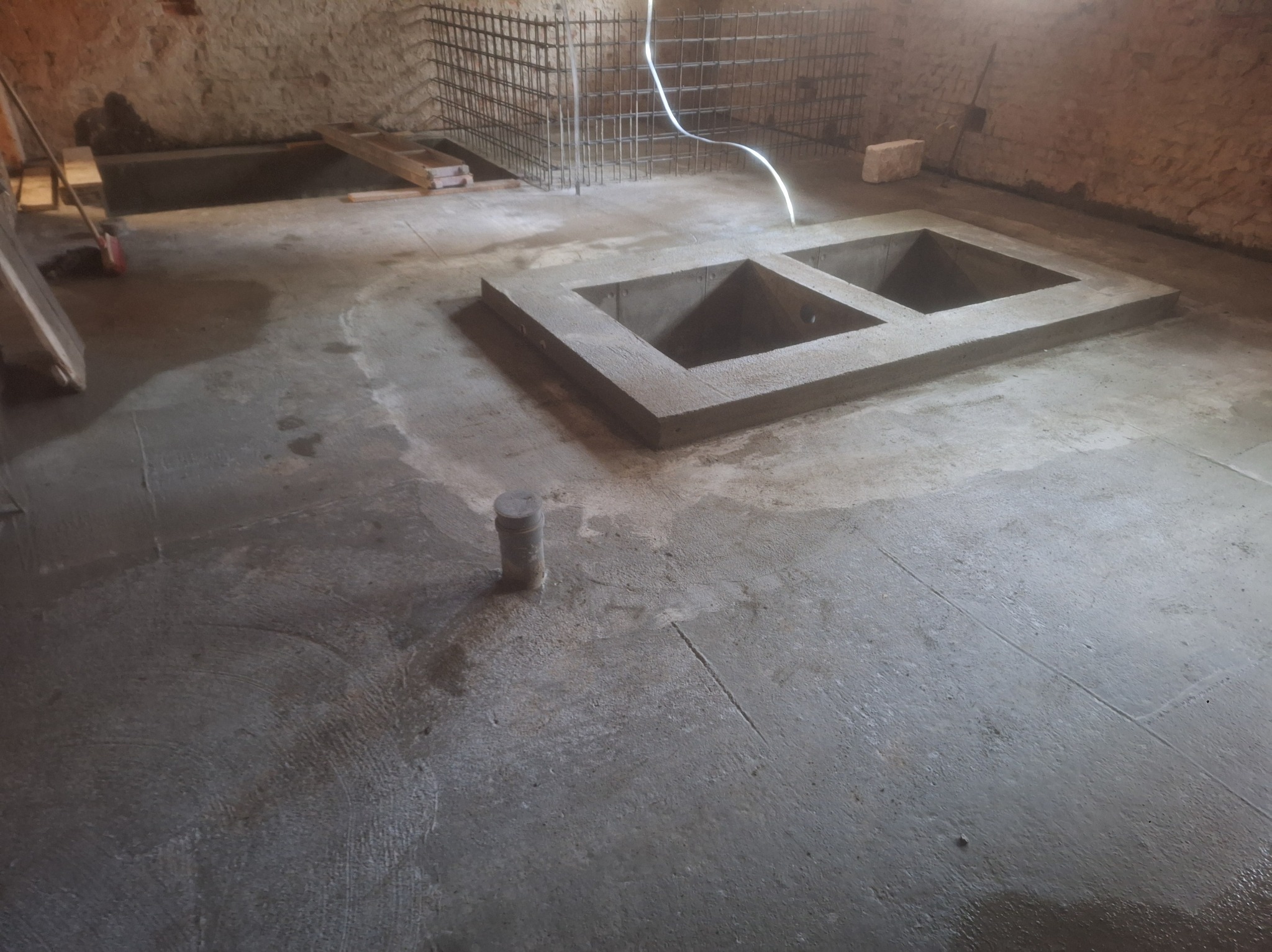 Surowy betonowy fundament z dwoma otworami w piwnicy, zbrojenie w tle. Widoczne ślady wilgoci na posadzce i surowe ściany. Etap budowy.