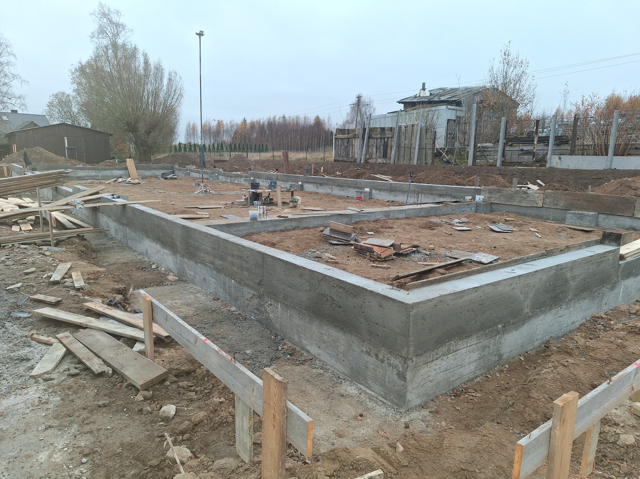 Zalany fundament pod budynek w Skrzeszewie Żukowskim. Widoczne szalunki i deski. Świeży beton, ziemia i drzewa w tle. Ujęcie z bliska, pochmurny dzień.