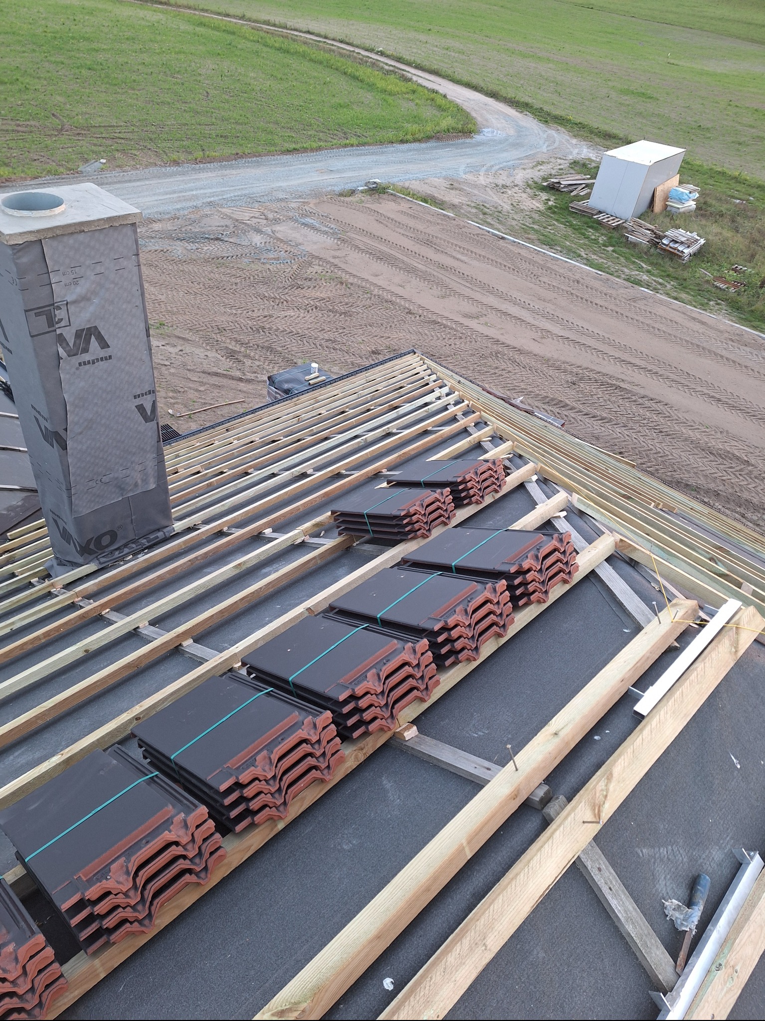 Układanie dachówki ceramicznej na konstrukcji więźby dachowej. Widoczny komin z izolacją i przygotowane pakiety dachówek. Tło: pole i droga gruntowa.