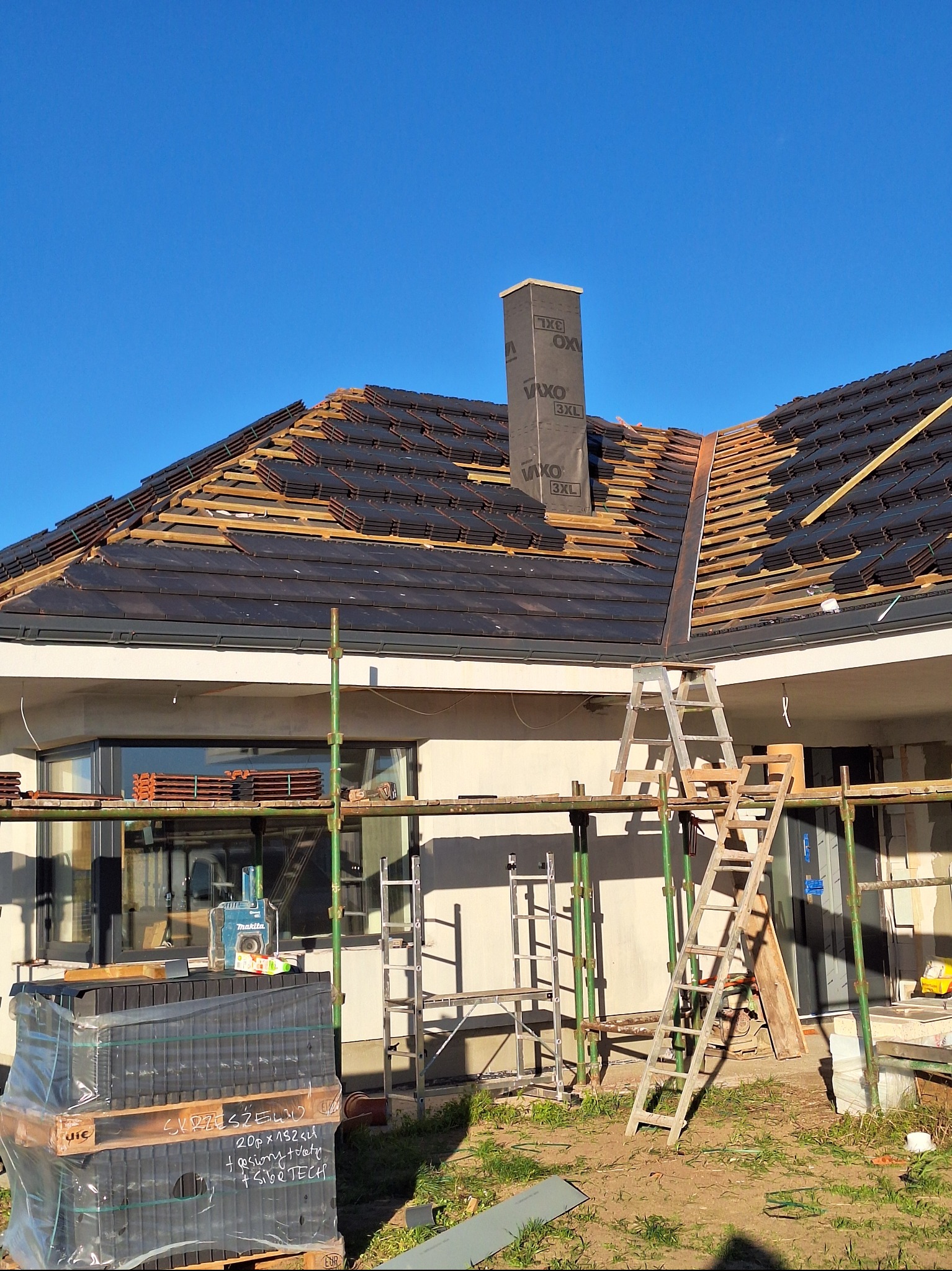 Układanie czarnej dachówki na dachu domu jednorodzinnego. Widoczne rusztowania, drabina i komin z czarną folią. Częściowo ułożony dach w słoneczny dzień.