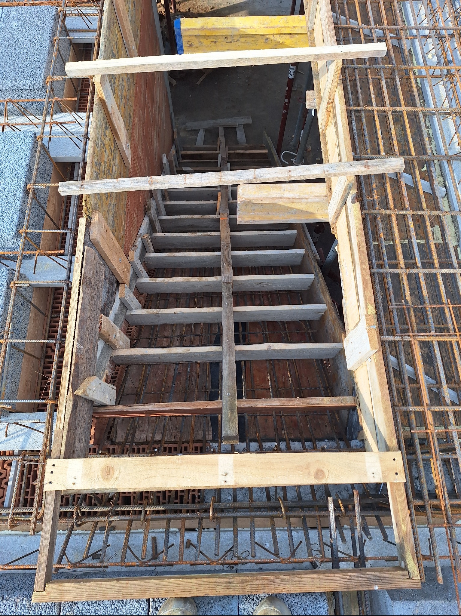 Wykonywanie schodów żelbetowych na budowie. Widok z góry na drewniany szalunek i zbrojenie. Elementy konstrukcyjne z cegły i betonu komórkowego w tle.