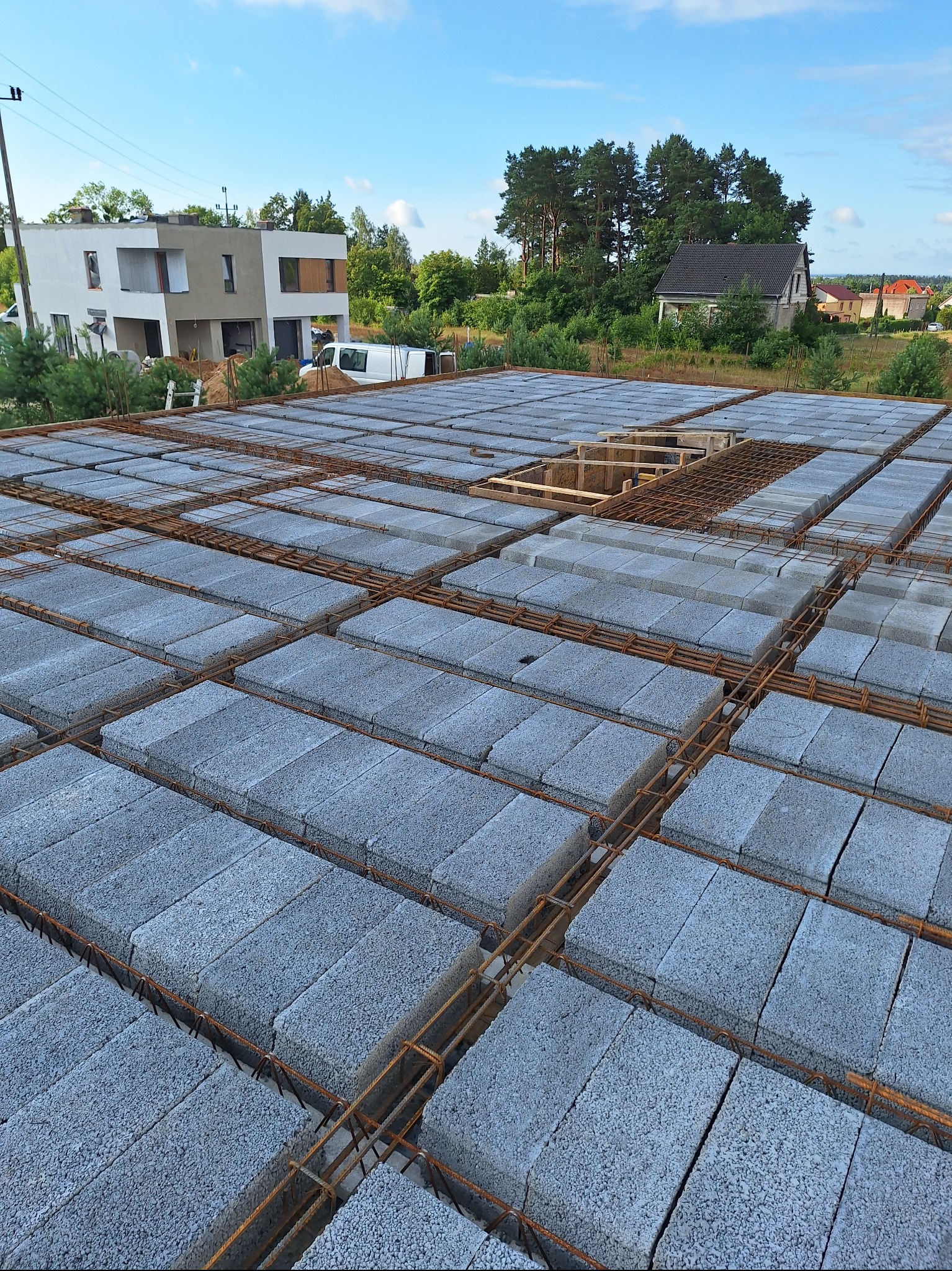 Wiązanie zbrojenia i układanie bloczków stropowych na budowie domu jednorodzinnego. Widok z góry na przygotowany strop, w tle budynek w stanie surowym otwartym.