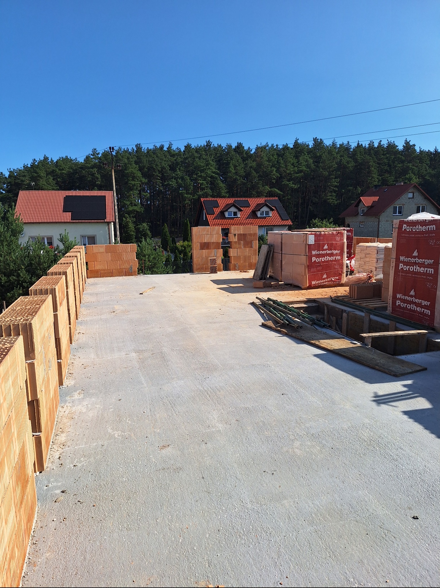 Budowa domu jednorodzinnego: widok z góry na fundamenty z cegły Porotherm, częściowo wzniesione ściany i materiały budowlane. W tle domy i las.