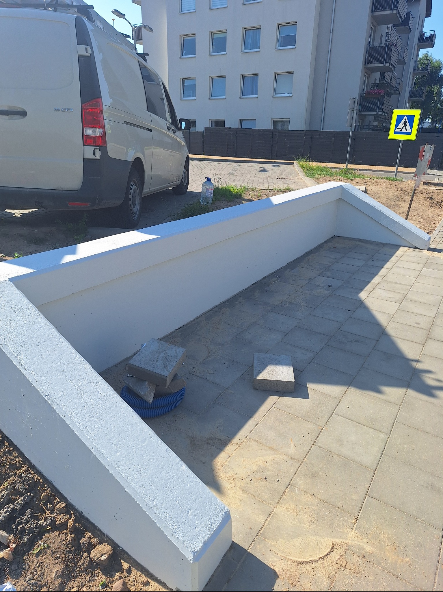 Świeżo pomalowana, biała betonowa konstrukcja obok chodnika z kostki brukowej, z zaparkowanym białym vanem w tle i znakiem przejścia dla pieszych.