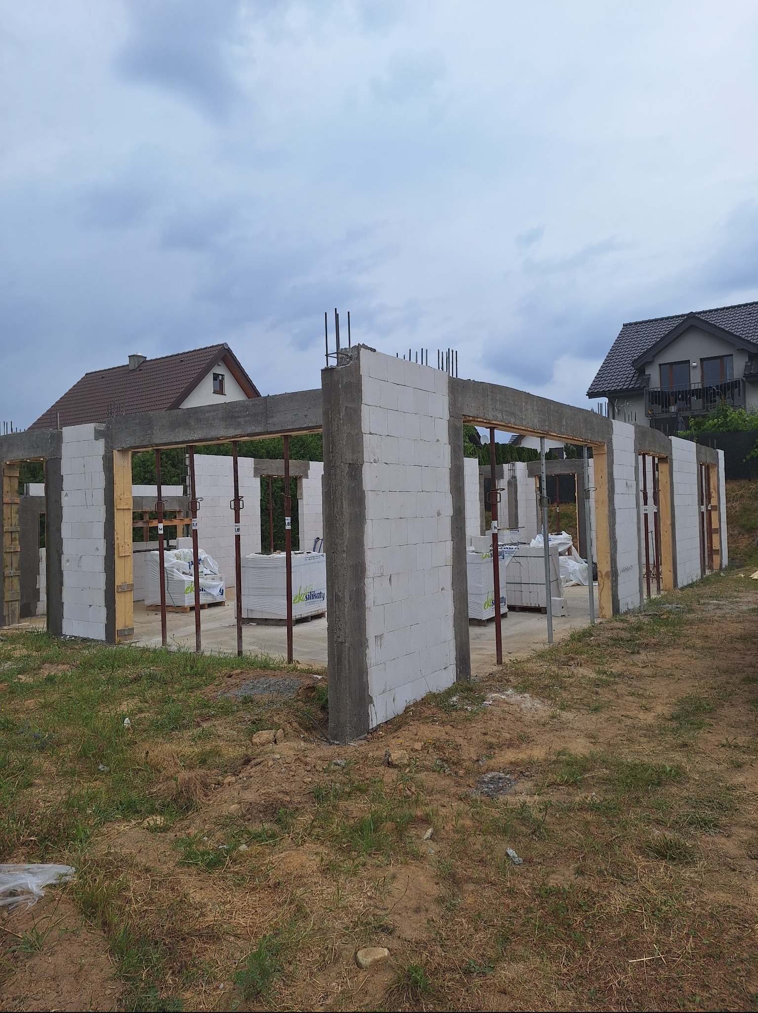 Wznoszenie ścian parteru domu z białych bloczków silikatowych, widoczne zbrojenia i podpory, w tle domy jednorodzinne pod pochmurnym niebem.