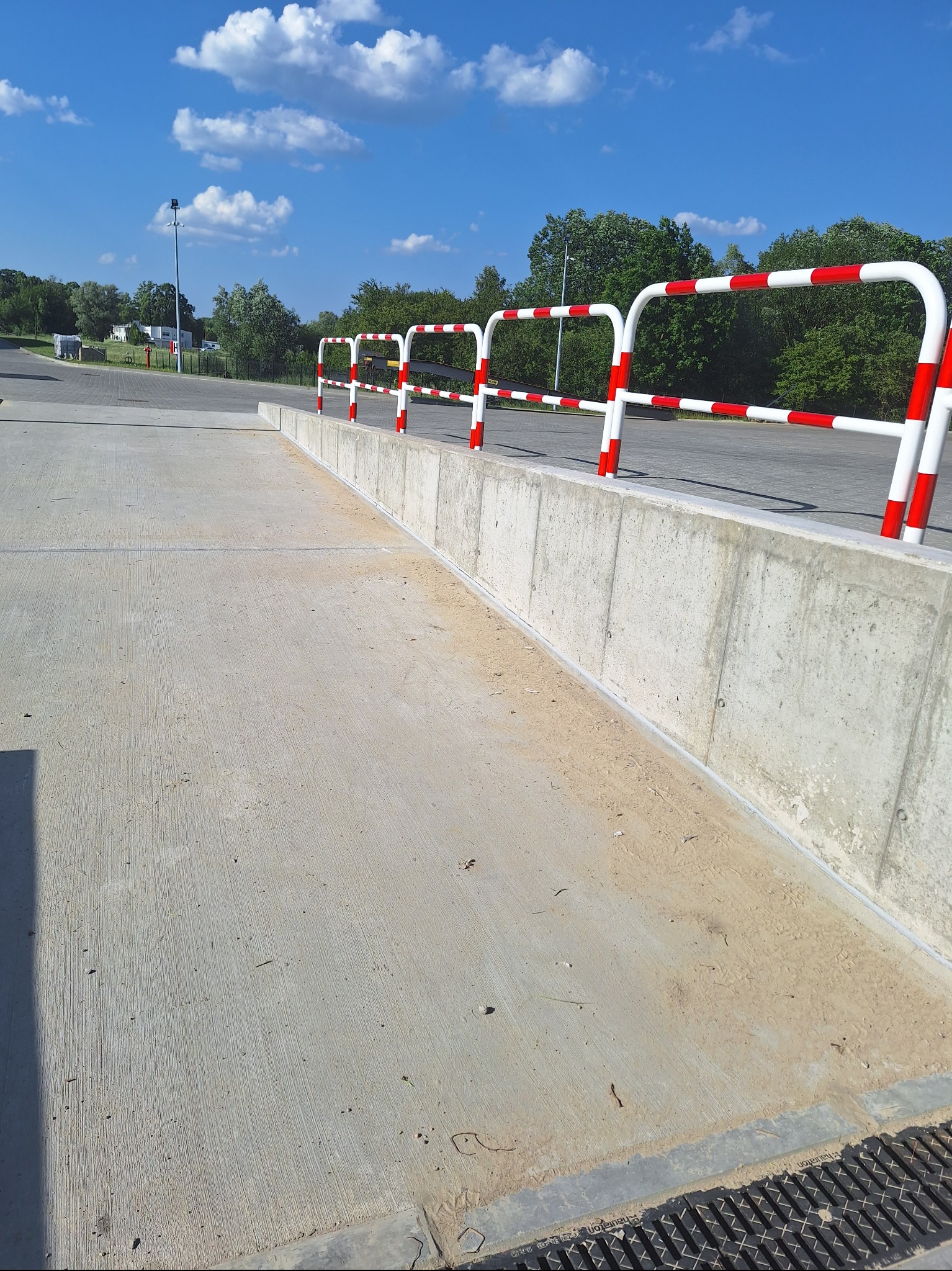 Betonowa rampa z barierkami w biało-czerwone pasy, prowadząca do parkingu. Widoczne drzewa i błękitne niebo z chmurami. Jasne, naturalne oświetlenie.
