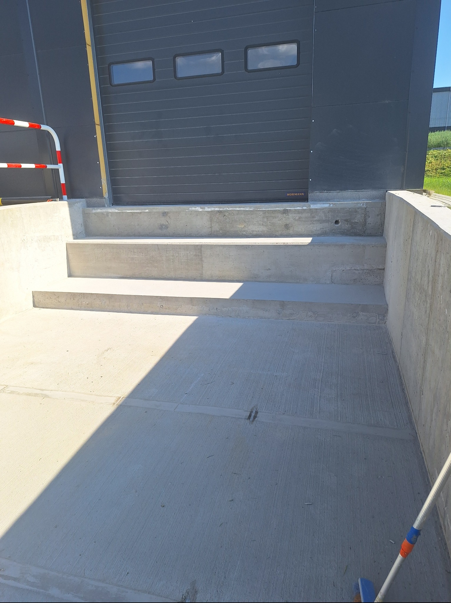 Betonowe schody wejściowe do budynku z szarą bramą segmentową i trzema oknami. Widoczny fragment barierki i cień. Ujęcie z dołu.