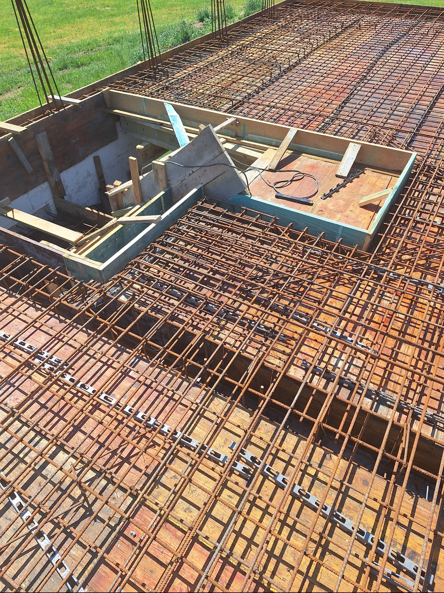 Zbrojenie płyty fundamentowej z widocznym otworem na instalacje, przygotowane do zalania betonem. Widoczne drewniane szalunki i elementy mocujące zbrojenie.