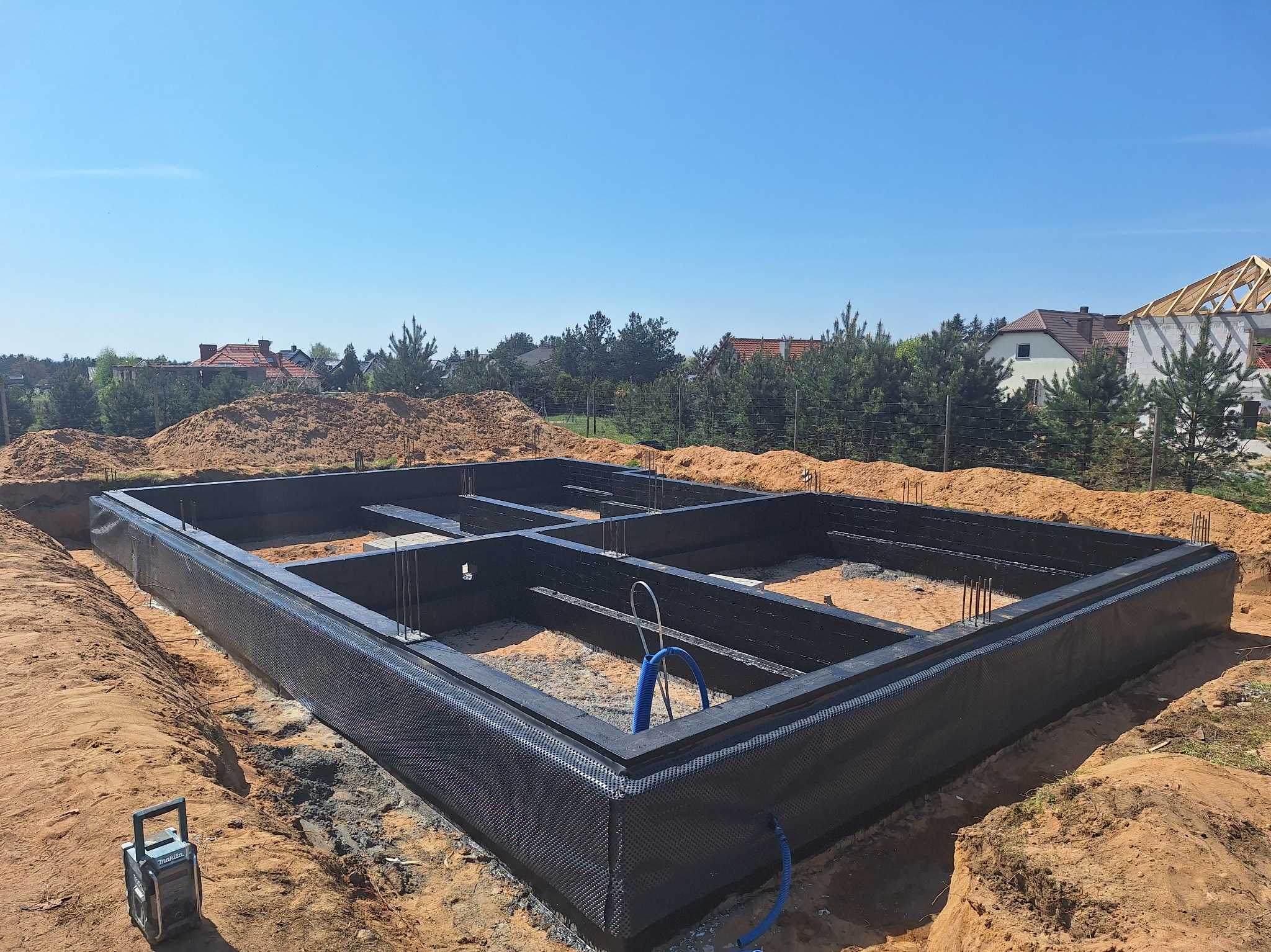 Zabezpieczone fundamenty domu jednorodzinnego z widoczną izolacją przeciwwilgociową, na tle wykopu i częściowo zalesionego terenu. Widoczny sprzęt budowlany.