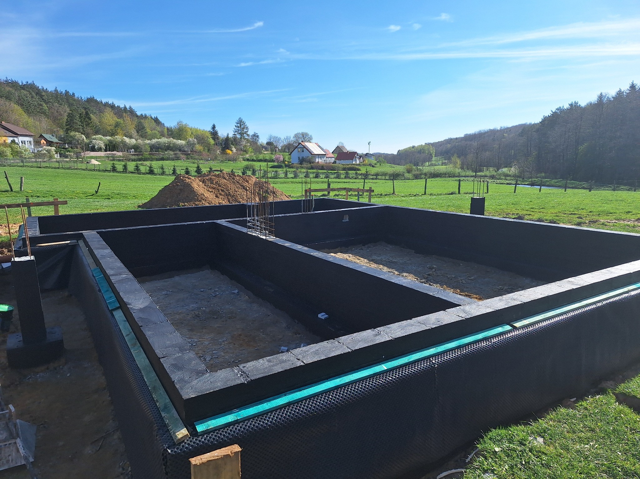 Fundamenty domu w trakcie budowy, z widoczną izolacją przeciwwilgociową i zbrojeniami, na tle zielonego krajobrazu z domami i lasem w oddali.