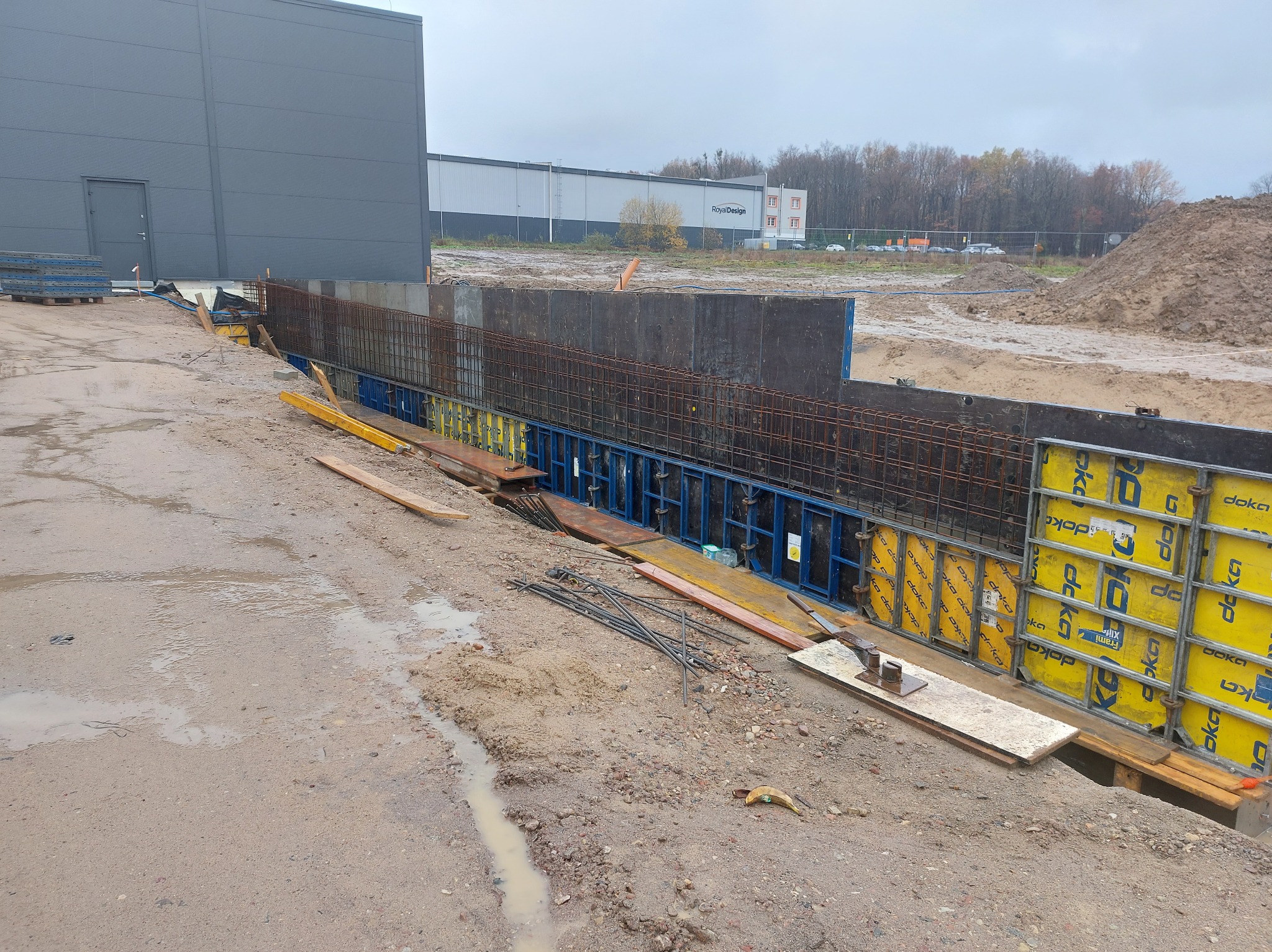Zbrojenie fundamentów w wykopie, szalunki Doka, widok na budynek przemysłowy w tle. Prace ziemne, przygotowanie do wylewania betonu. Detal: żółte szalunki.