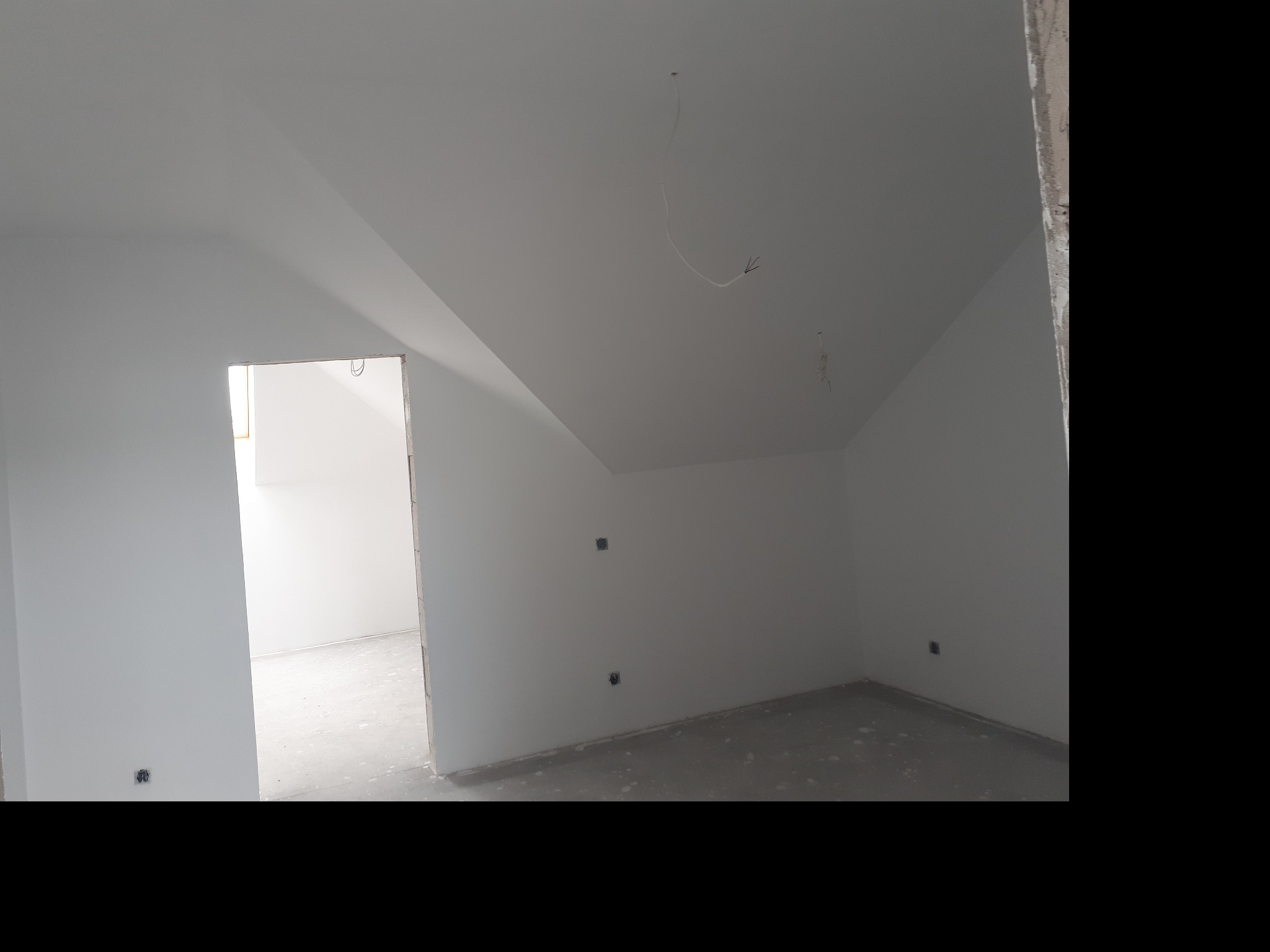 Wnętrze poddasza po malowaniu na biało, widoczne gniazdka elektryczne, surowy beton na podłodze i wystające kable. Otwór drzwiowy prowadzi do kolejnego pomieszczenia.