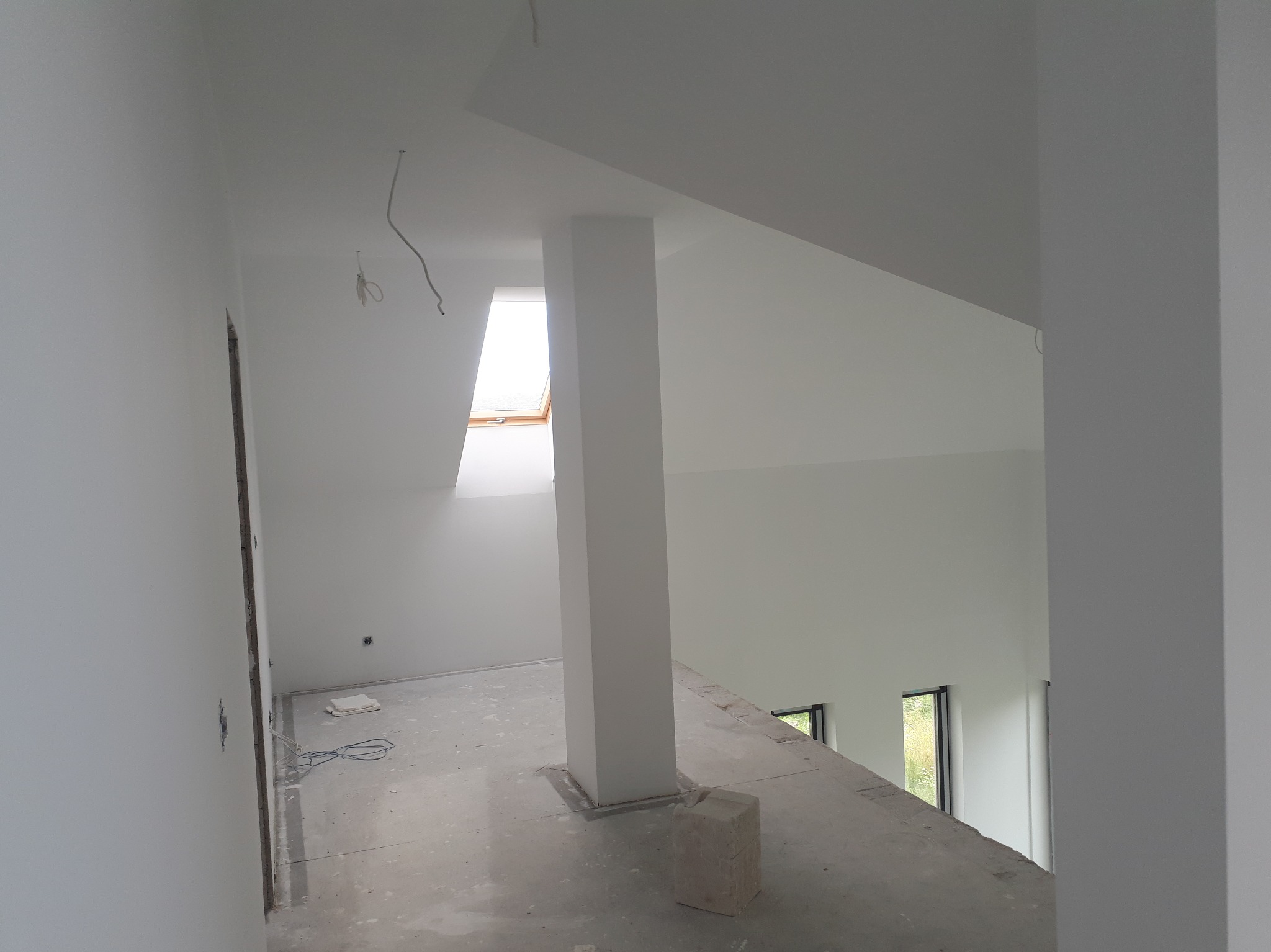 Białe, świeżo pomalowane poddasze z oknem dachowym i filarem. Widoczne kable elektryczne i surowa podłoga. Minimalistyczne wnętrze w trakcie wykańczania.