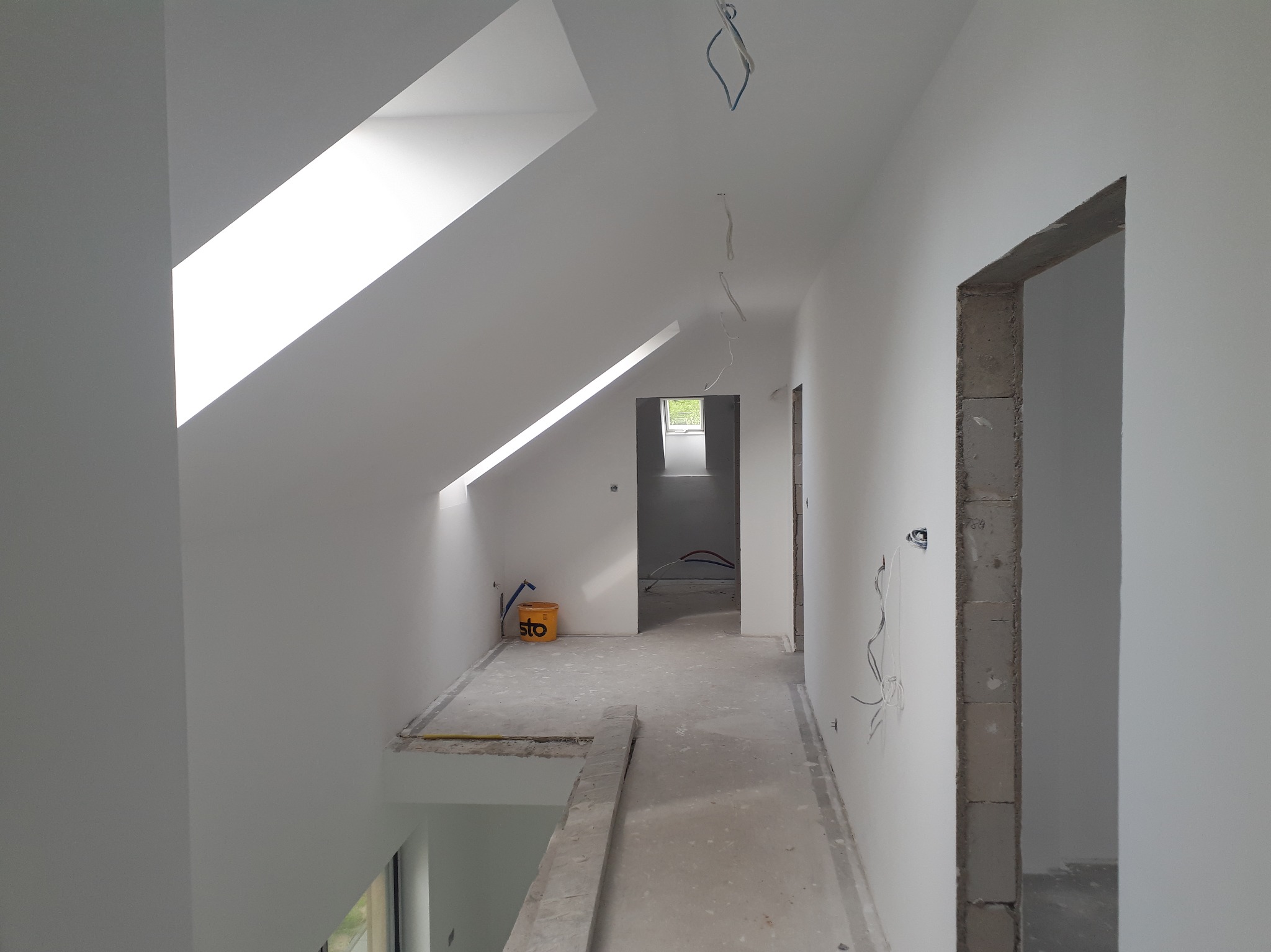 Poddasze po malowaniu na biało, widoczne okno dachowe, surowa ościeżnica i wystające przewody elektryczne. Na podłodze stoi wiadro farby.