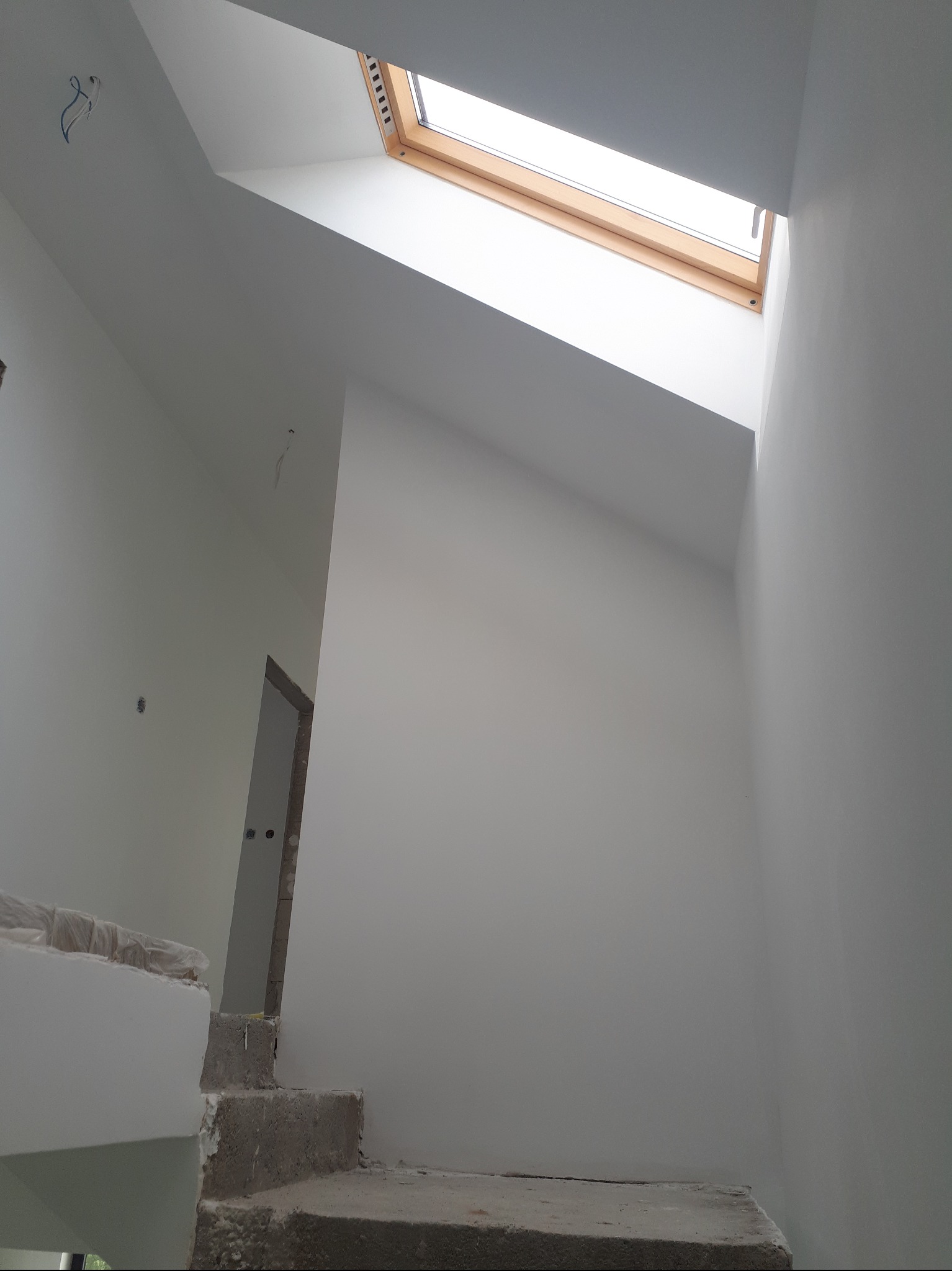 Wnętrze poddasza po malowaniu na biało, widoczne schody w trakcie wykończenia i okno dachowe z drewnianą ramą. Minimalistyczny design, surowe wykończenie.