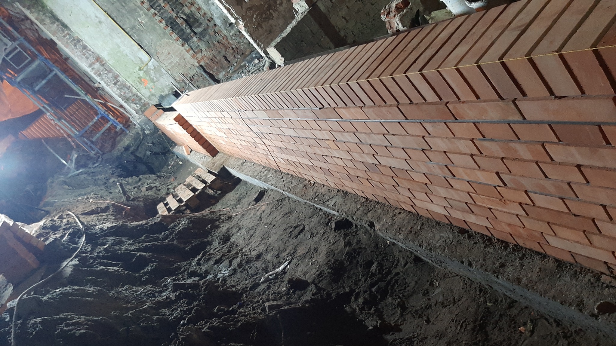 Wznoszenie muru z cegły klinkierowej w wykopie fundamentowym, widoczne zbrojenie i fragment rusztowania. Ujęcie z dołu, podkreślające pionowość konstrukcji.