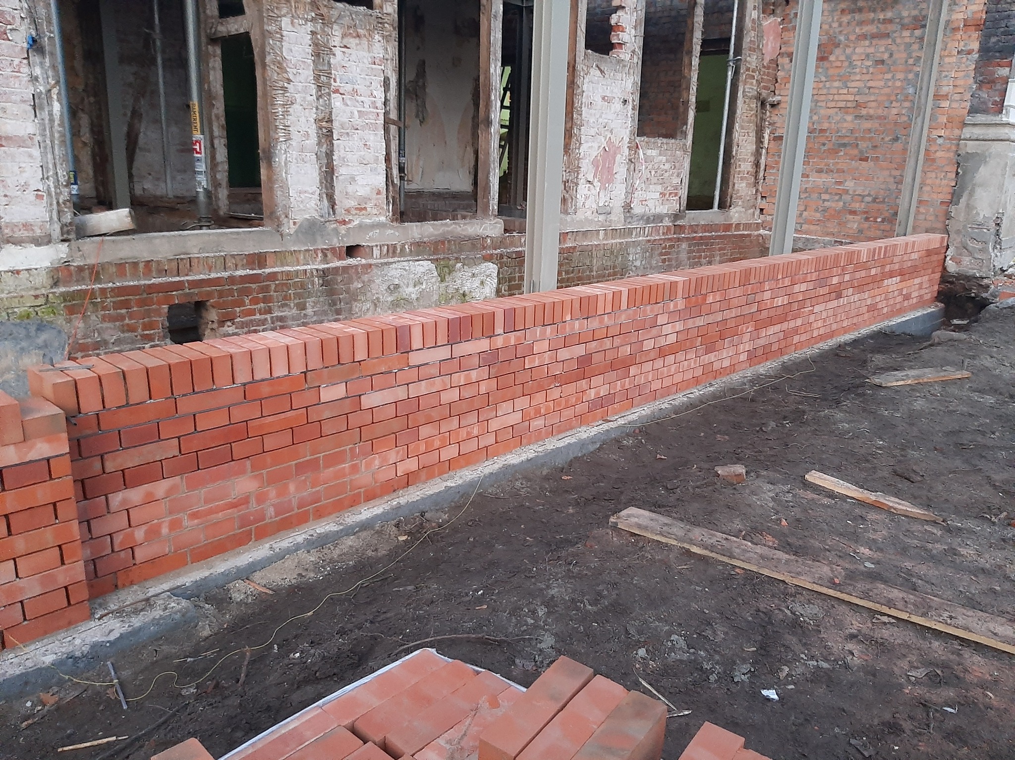 Ceglana ściana w trakcie budowy, z widocznymi fragmentami starej konstrukcji w tle. Surowy teren budowy z ziemią i deskami. Czerwona cegła kontrastuje z szarością otoczenia.