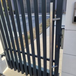 PPHU DECOR Ogrodzenia Łukasz Orynek - Nowoczesna, antracytowa furtka z pionowych profili, z srebrną klamką i zamkiem, zamontowana przy jasnym murze. Widoczny fragment posesji w tle.