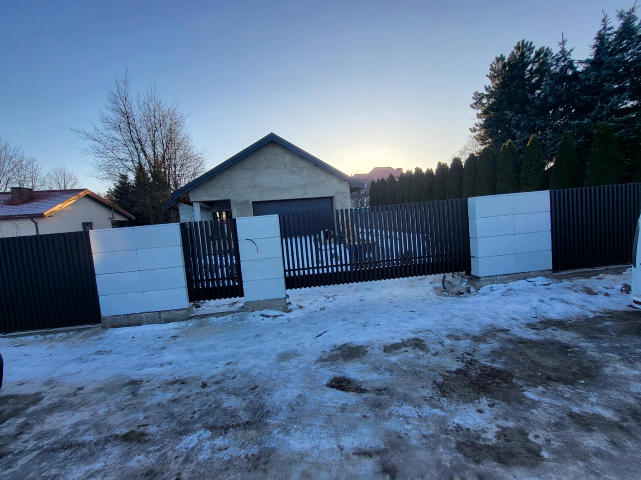 Nowoczesne ogrodzenie z pionowych, grafitowych paneli i białych, kwadratowych słupków na tle zimowego krajobrazu i domu z garażem.