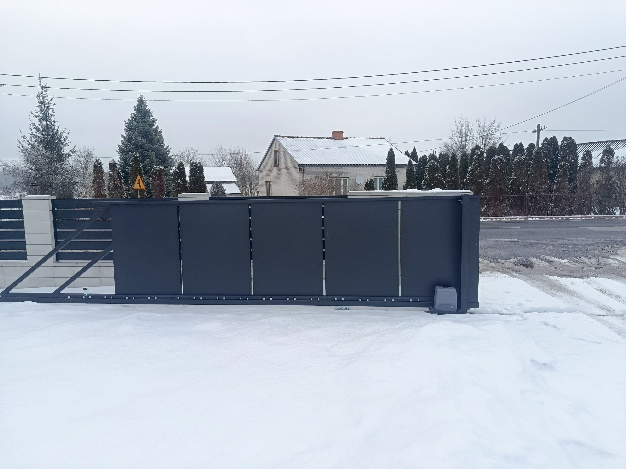 Nowoczesna, przesuwna brama wjazdowa w kolorze grafitowym, z widocznym napędem, na tle zimowego krajobrazu z domem i drzewami. Minimalistyczny design.