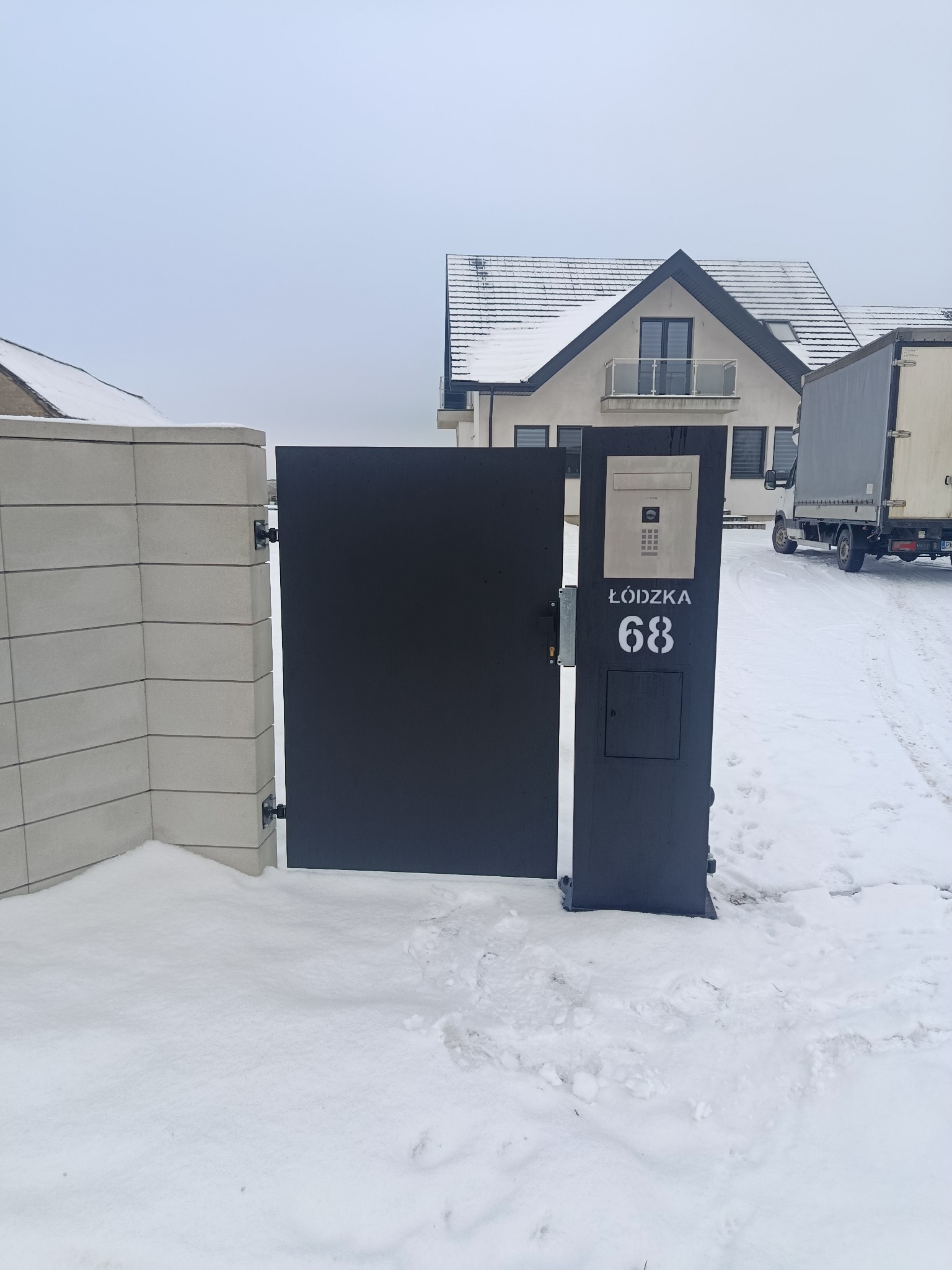 Nowoczesna, grafitowa brama wjazdowa z numerem domu 'Łódzka 68' i domofonem, na tle ośnieżonego podwórza i domu. Minimalistyczny design.