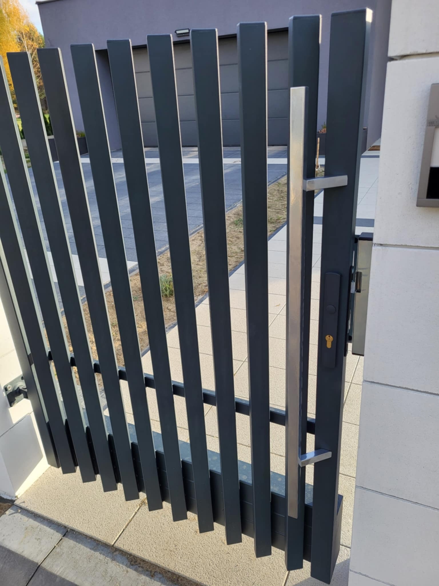 Nowoczesna, antracytowa furtka z pionowych profili, z srebrną klamką i zamkiem, zamontowana przy jasnym murze. Widoczny fragment posesji w tle.