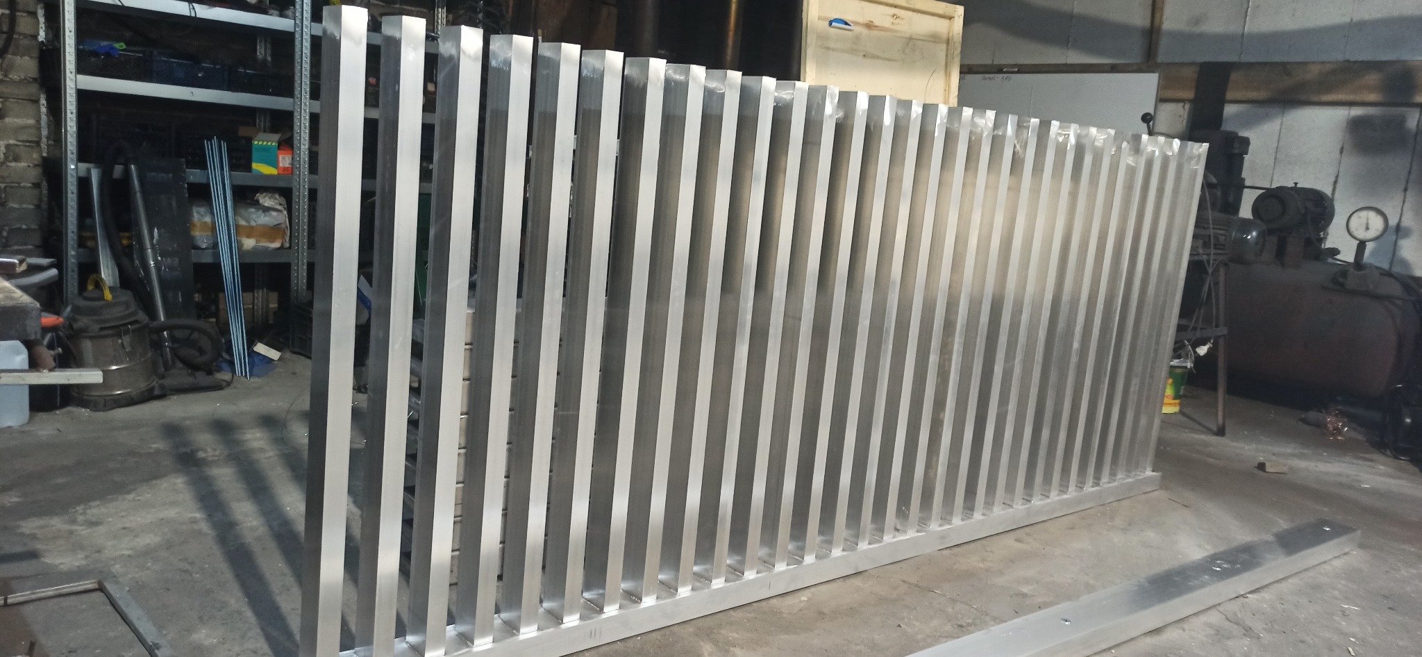 Aluminiowe ogrodzenie lamelowe w warsztacie, widoczne pionowe profile i rama. W tle narzędzia i sprzęt warsztatowy. Surowy, industrialny charakter.
