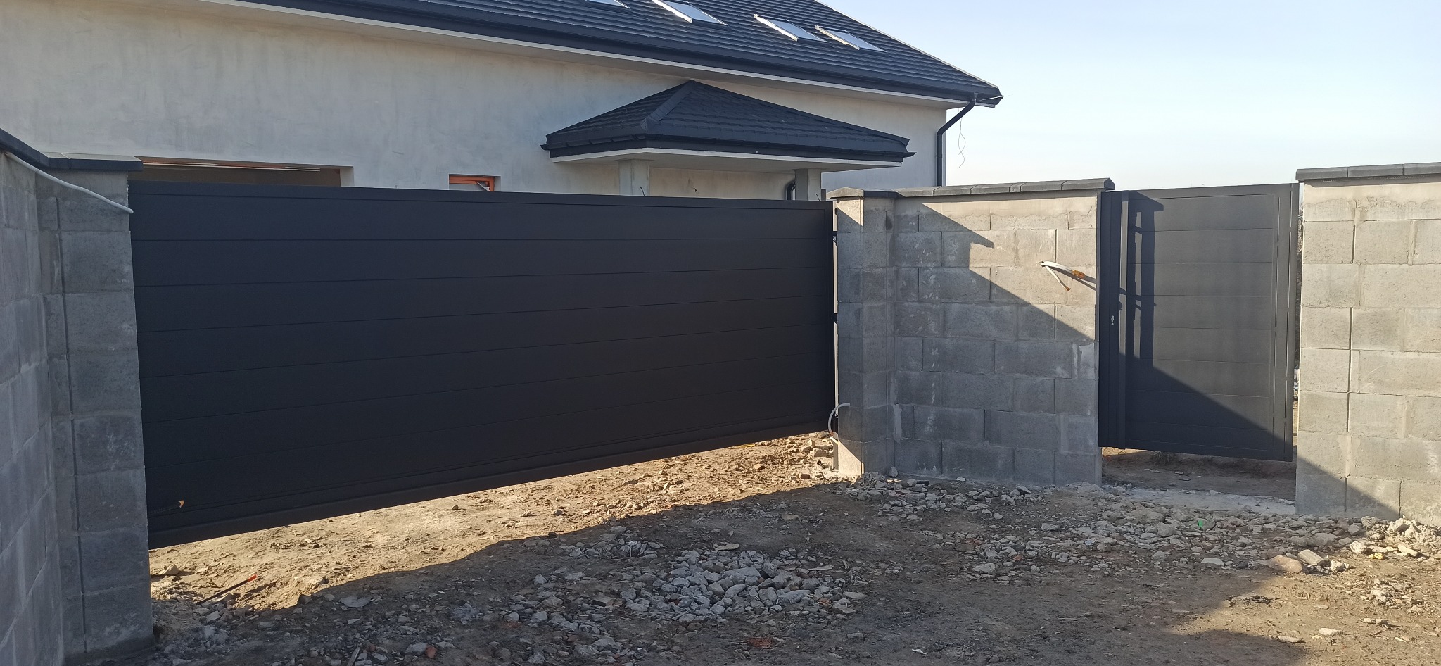 Nowoczesna, przesuwna brama wjazdowa w kolorze antracytowym, zamontowana na betonowych słupkach. Widoczne detale wykończenia i surowy teren wokół.