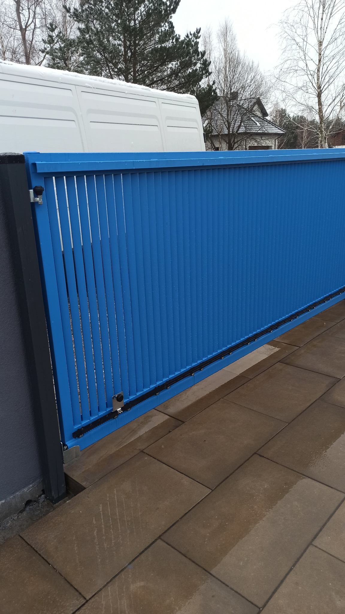 Niebieska brama przesuwna z pionowymi panelami, widoczna z boku, z fragmentem białego samochodu dostawczego w tle i mokrą kostką brukową na pierwszym planie.