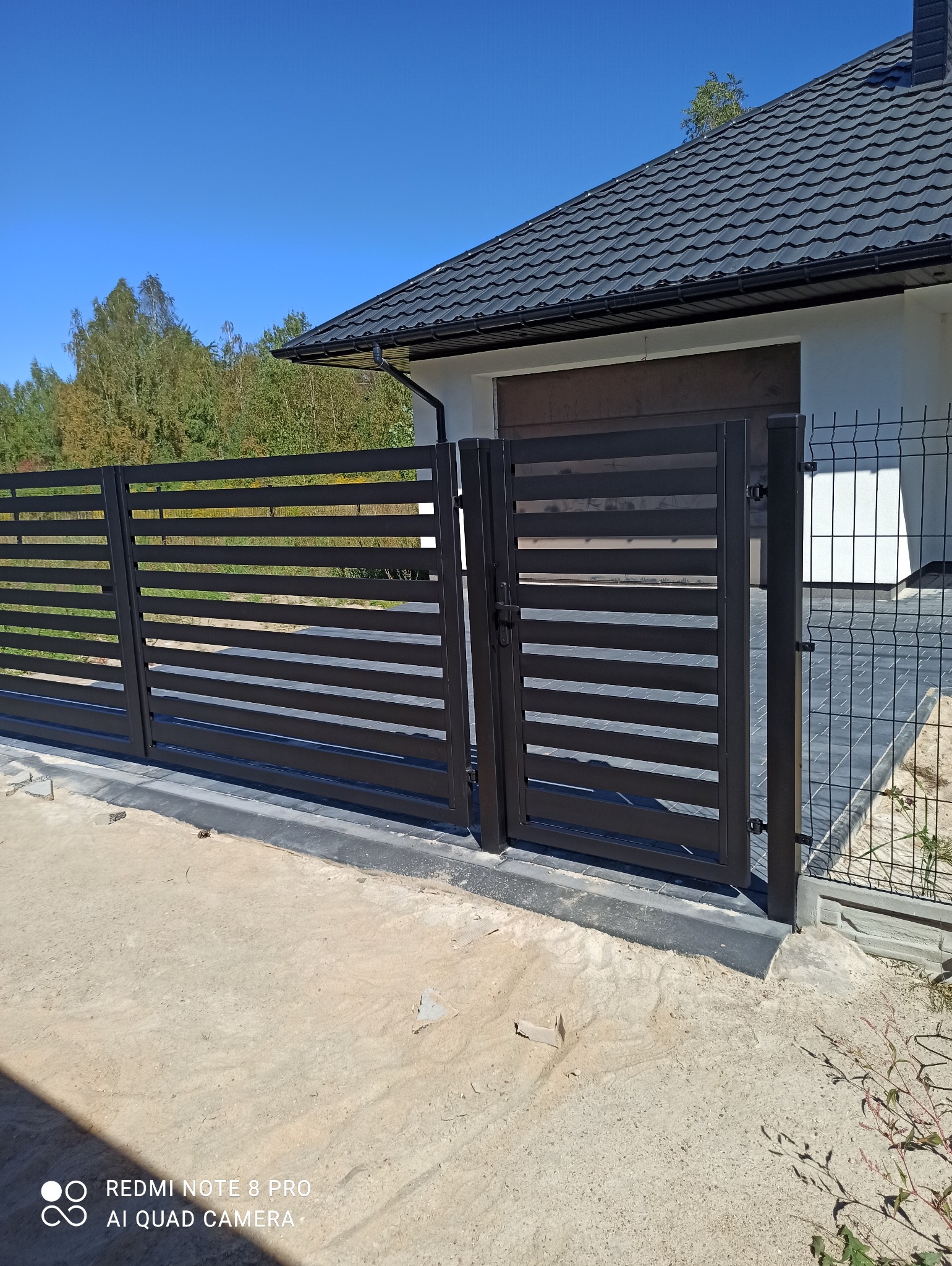 Nowoczesna, pozioma brama przesuwna w kolorze antracytowym, z widoczną furtką i fragmentem ogrodzenia panelowego. Tło stanowi dom z ciemną dachówką i błękitne niebo.