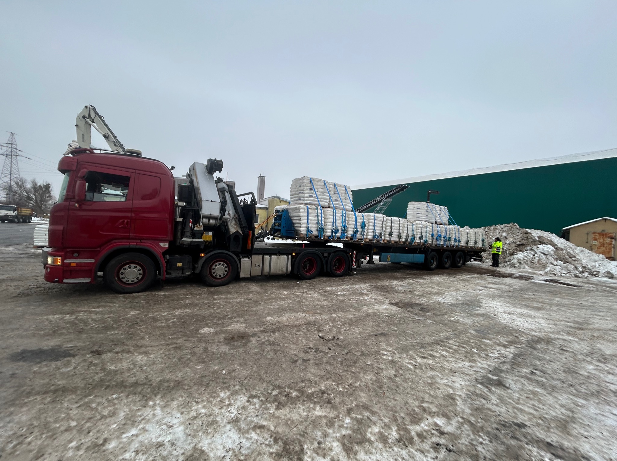 Czerwona ciężarówka z dźwigiem przewozi palety z workami na przyczepie. W tle zielony budynek i pracownik w odblaskowej kamizelce. Zimowy krajobraz.