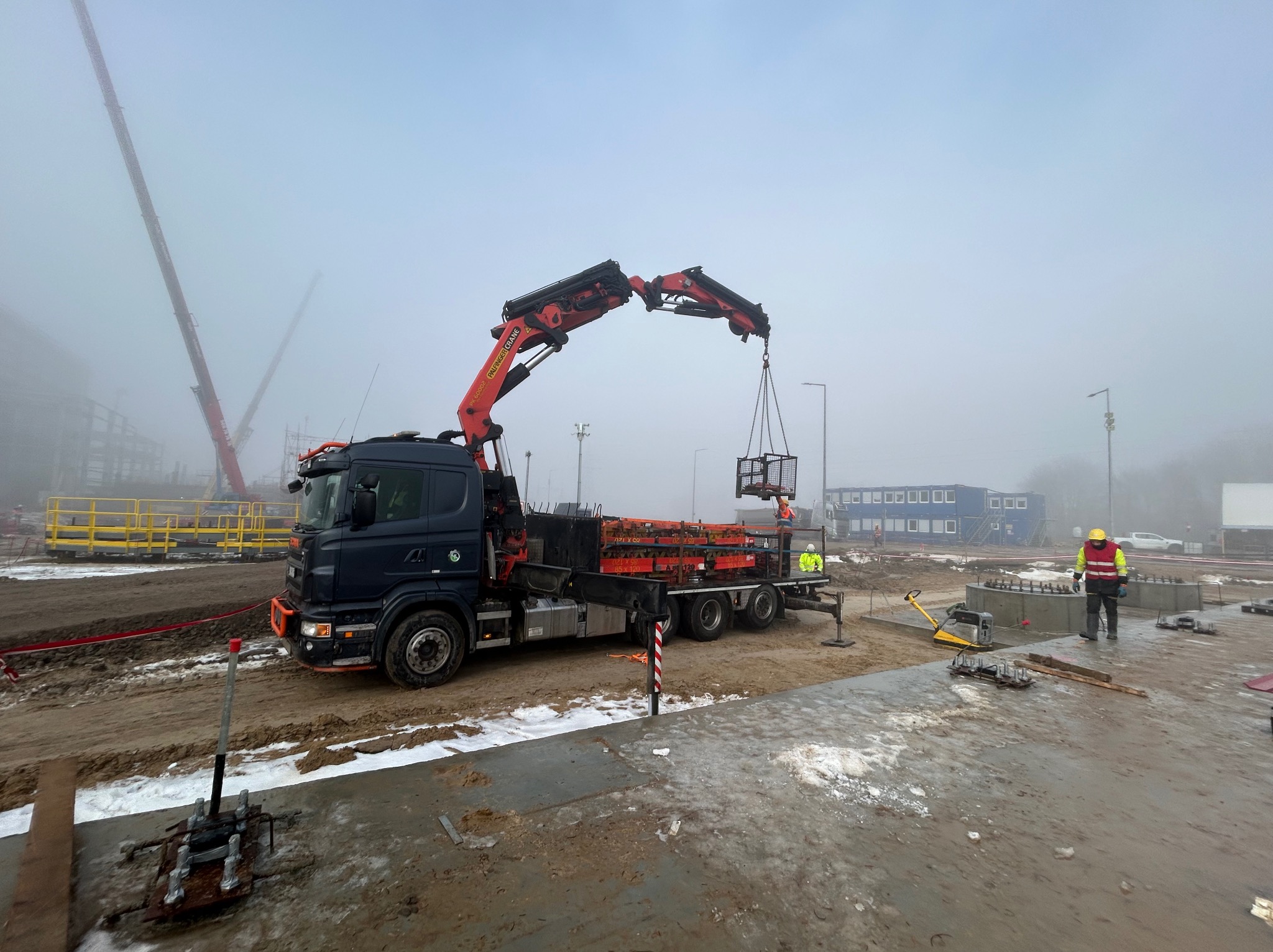 Dźwig samochodowy z wysięgnikiem transportuje ładunek na budowie w Elblągu. W tle zamglony krajobraz i konstrukcja w trakcie budowy. Praca na wysokości.