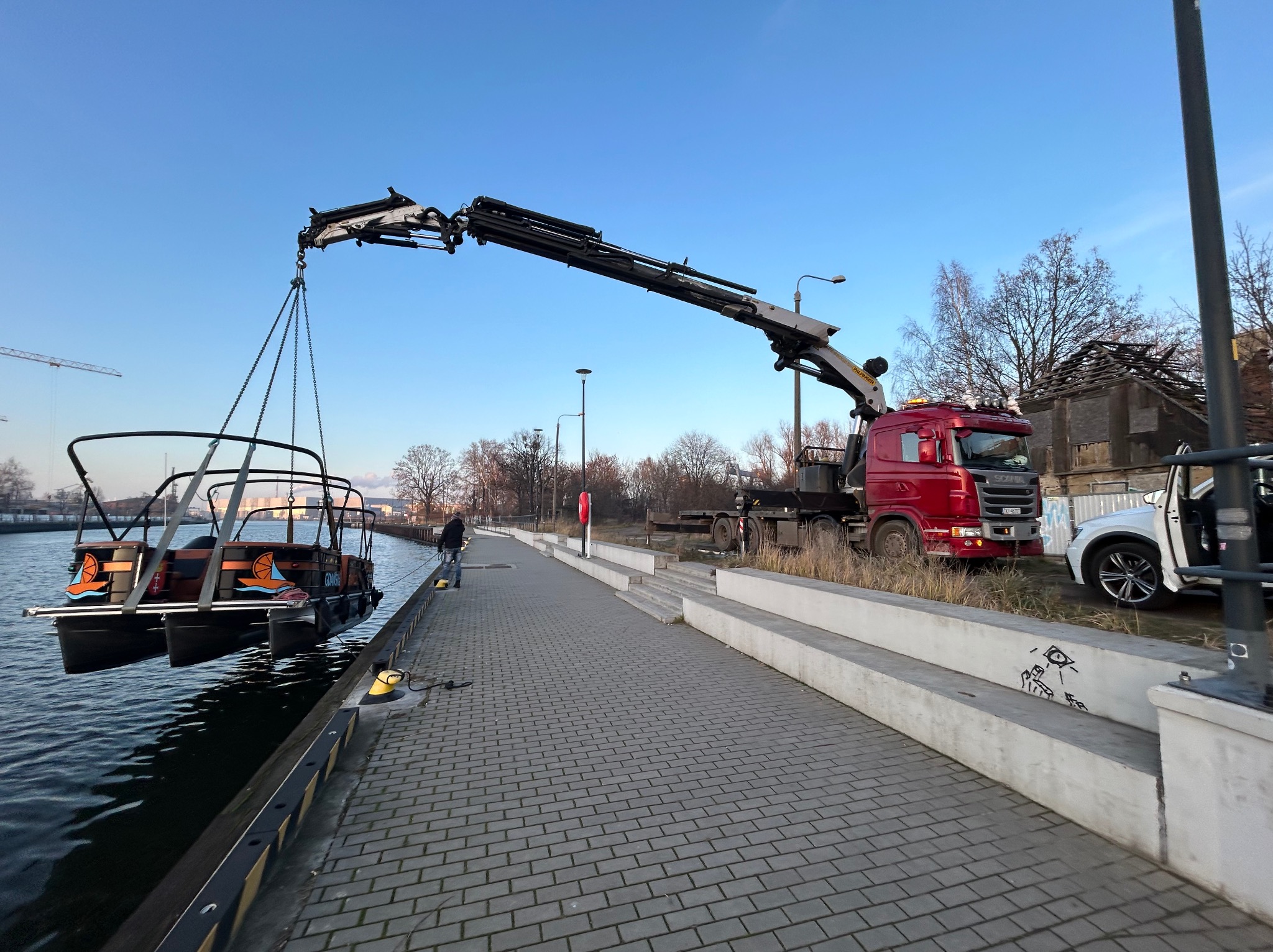Czerwony dźwig samochodowy podnosi ponton z napisem Gdańsk nad wodą. Operator w pobliżu. Brukowane nabrzeże i budynki w tle.