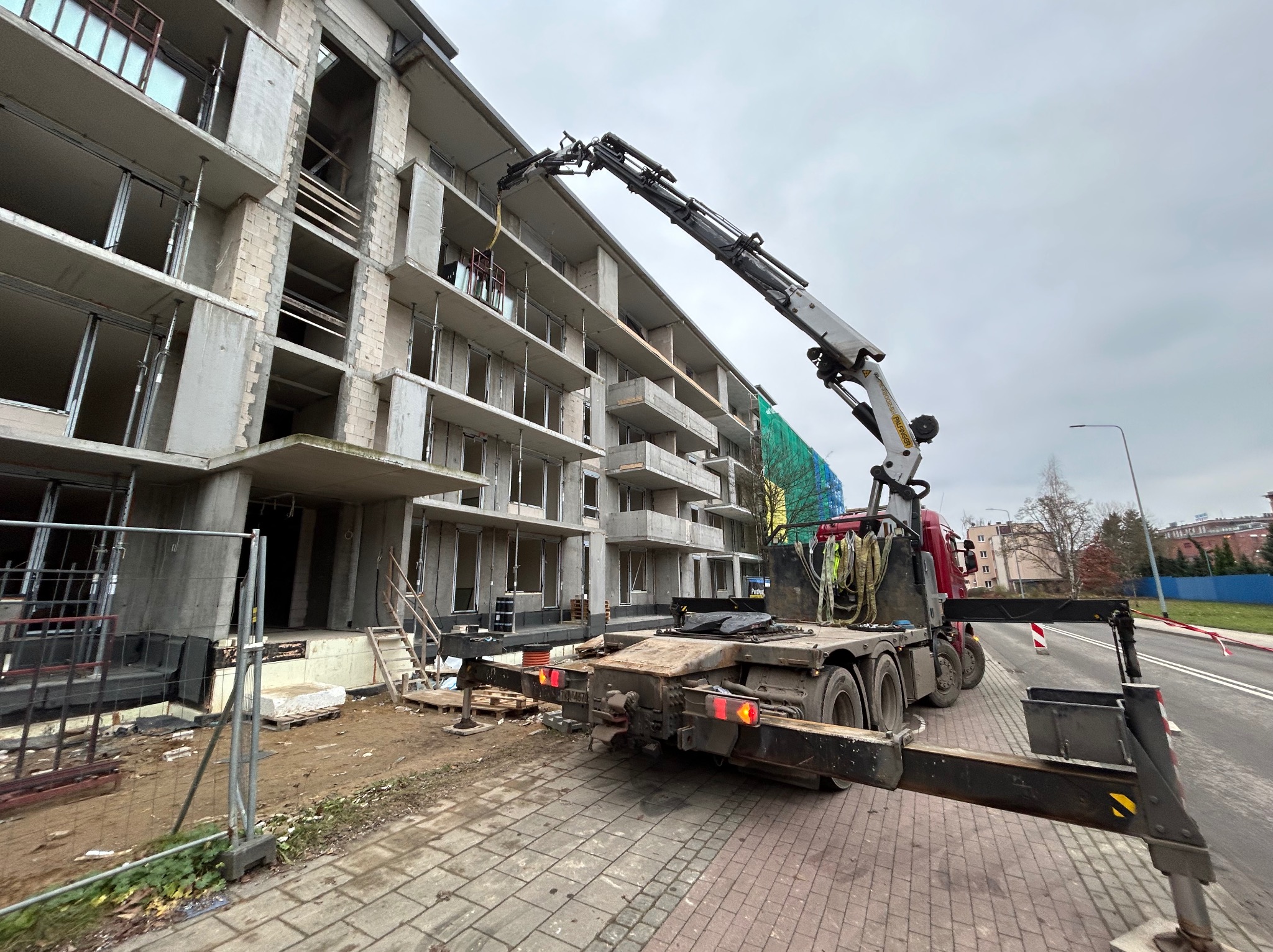 Dźwig samochodowy podnosi element na budowę wielopiętrowego budynku z betonu komórkowego. Widok z dołu, pochmurne niebo w tle.