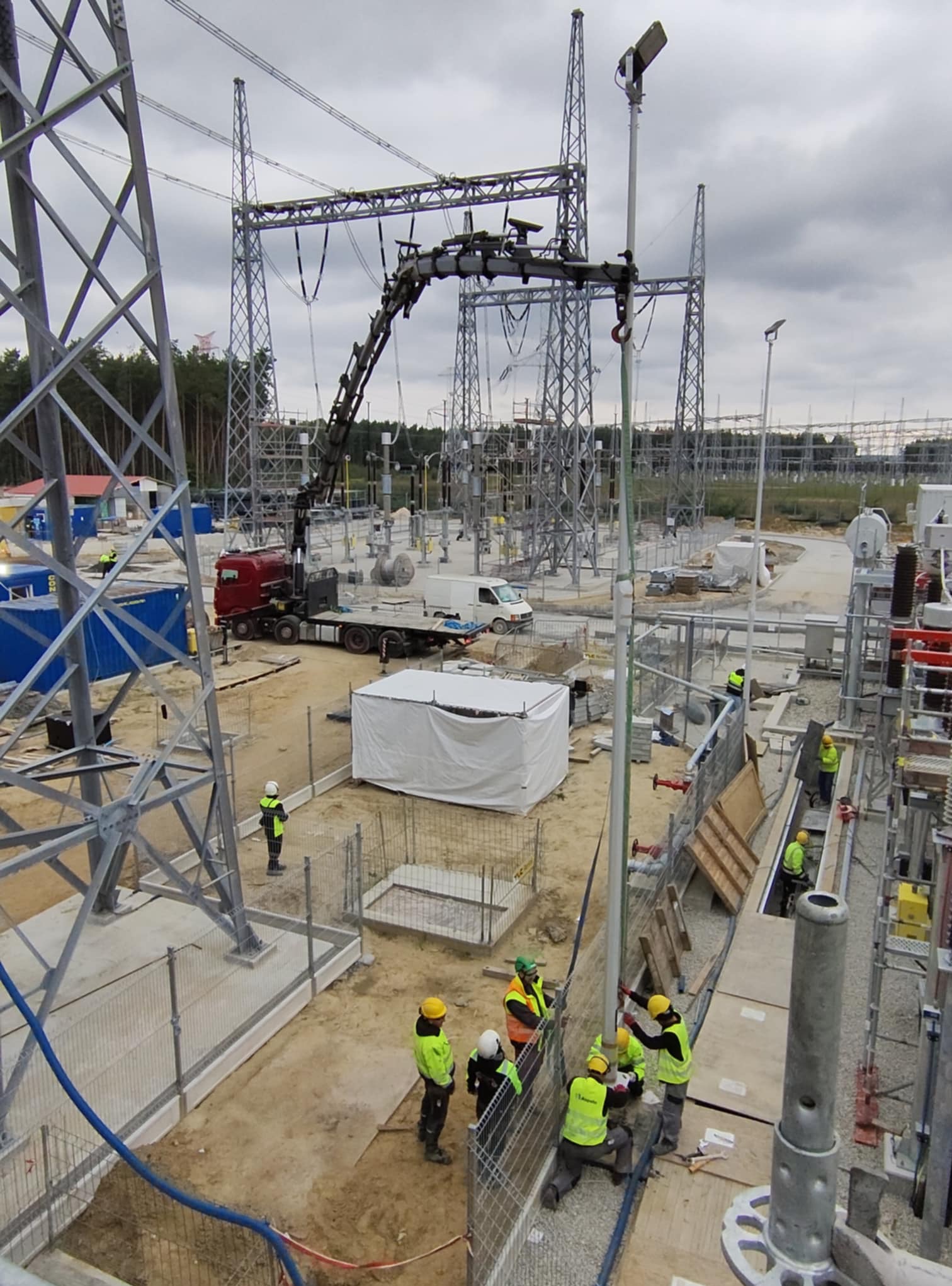 Montaż konstrukcji stalowej na terenie stacji transformatorowej. Widoczny dźwig samochodowy i pracownicy w kaskach. W tle słupy wysokiego napięcia i las.