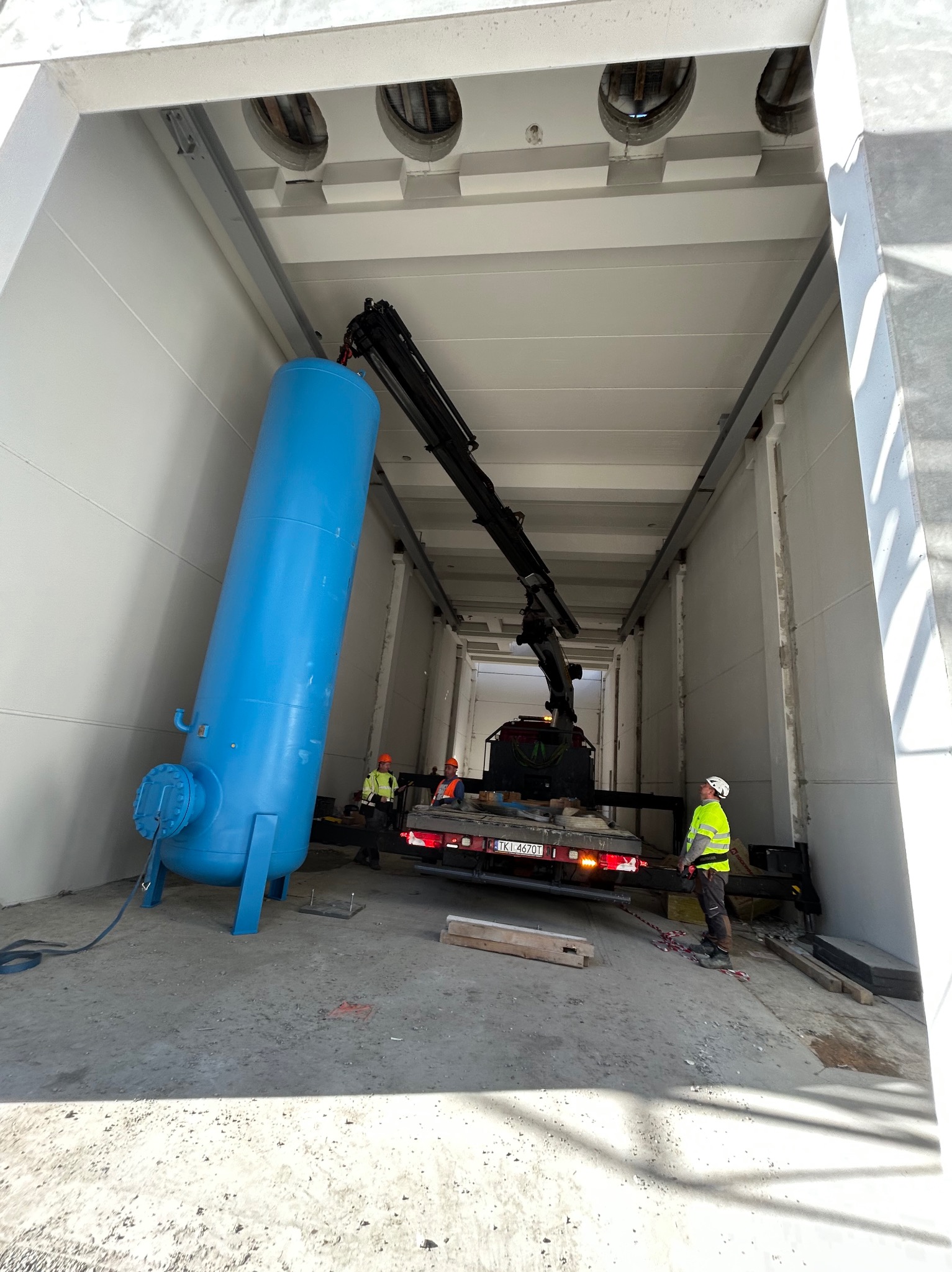 Dźwig unosi błękitny zbiornik wewnątrz hali o białych ścianach. Pracownicy w odblaskowych ubraniach nadzorują operację. Widoczna tablica rejestracyjna pojazdu.