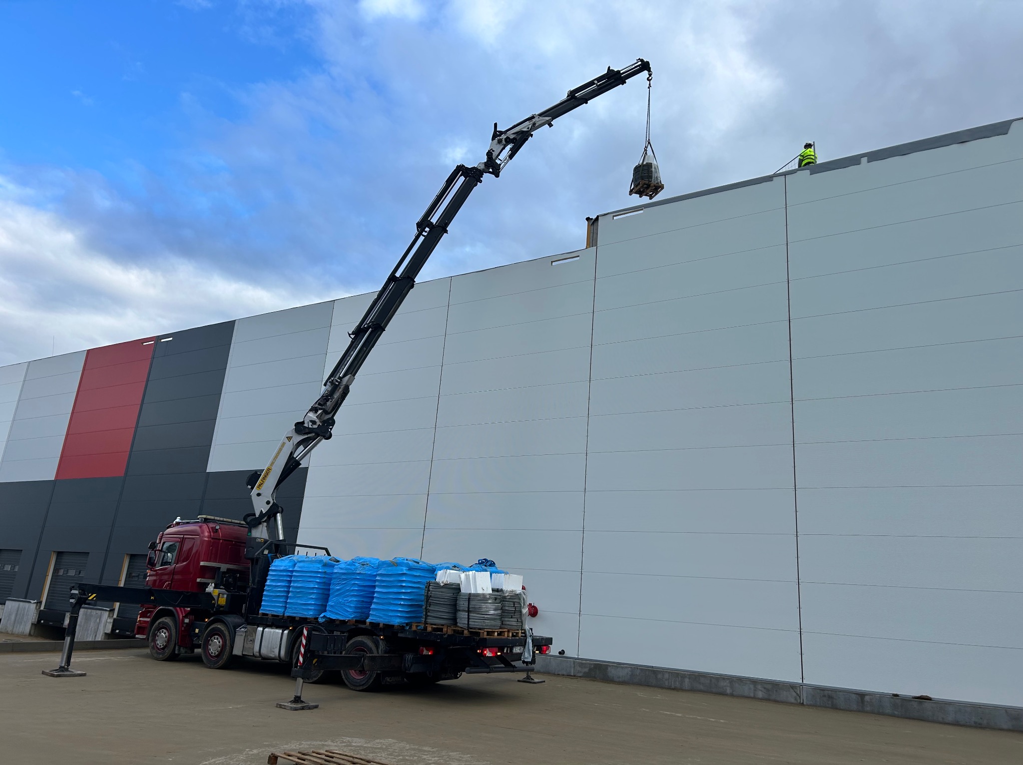 Czerwony dźwig samochodowy z ładunkiem na palecie, podnosi materiały na dach hali magazynowej. Pracownik w żółtej kamizelce na dachu. Błękitne niebo w tle.