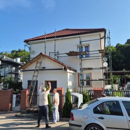 Jaroslaw Rudzki - Dwóch mężczyzn przy rusztowaniu maluje elewację domu jednorodzinnego w słoneczny dzień. Widoczna drabina i samochód osobowy. Prace w toku.