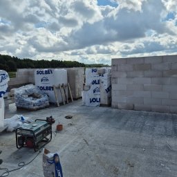 Jaroslaw Rudzki - Wznoszenie muru z bloczków Solbet na budowie. Widoczne palety z materiałami, agregat prądotwórczy i worki z zaprawą. W tle las i błękitne niebo z chmurami.